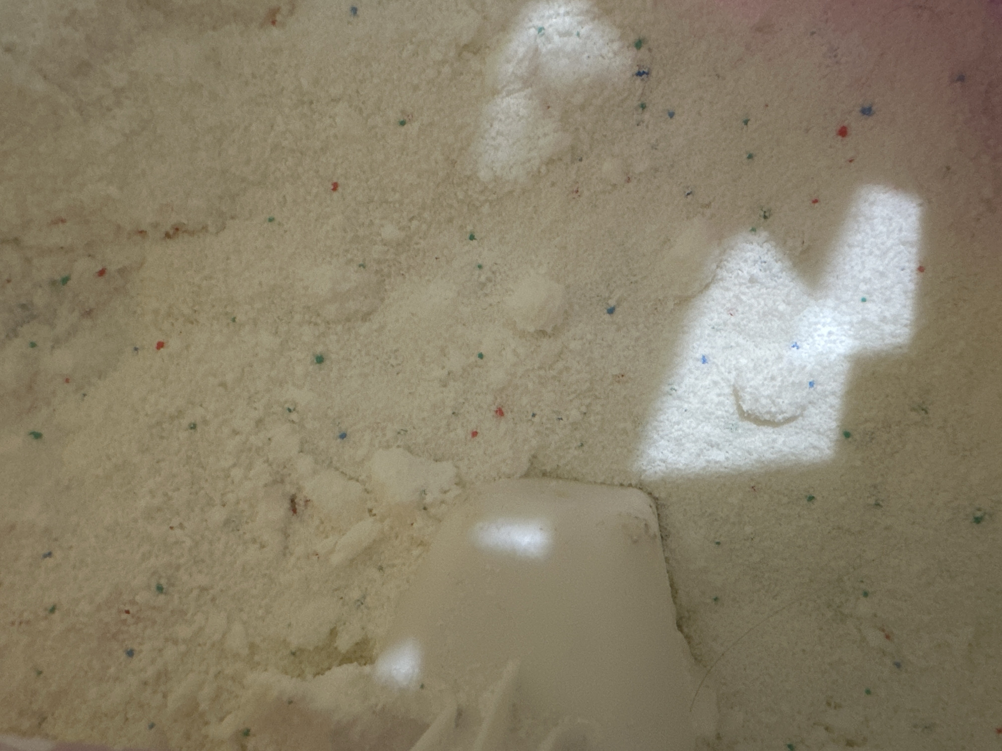 This is a close-up, overhead shot of powdered detergent with small colored beads interspersed throughout. The detergent is off-white and appears to be somewhat clumpy in places. A white scoop, likely plastic, is visible on the left side of the frame, partially submerged in the powder. Shadows from an unseen light source create bright highlights and darker areas on the surface of the detergent. The image does not contain any contextual information about a specific location in Birobidzhan, Russia, nor does it depict any people, actions, or interactions.