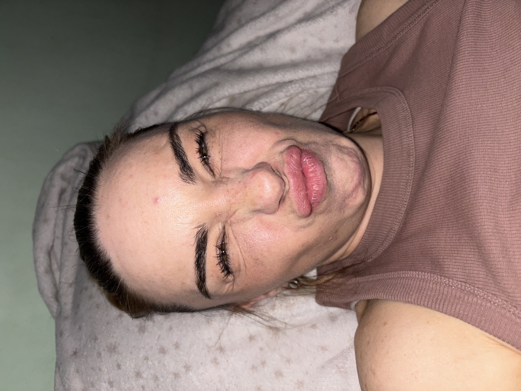 The image shows a close-up, overhead view of a young woman lying down, making a fish-face. She is wearing a brown ribbed tank top. Her face is the central focus, with her eyes squinted and her lips puckered outwards. The background consists of a textured white blanket and a muted green wall. No specific location in the city of Unknown, Russia, can be identified from the image.