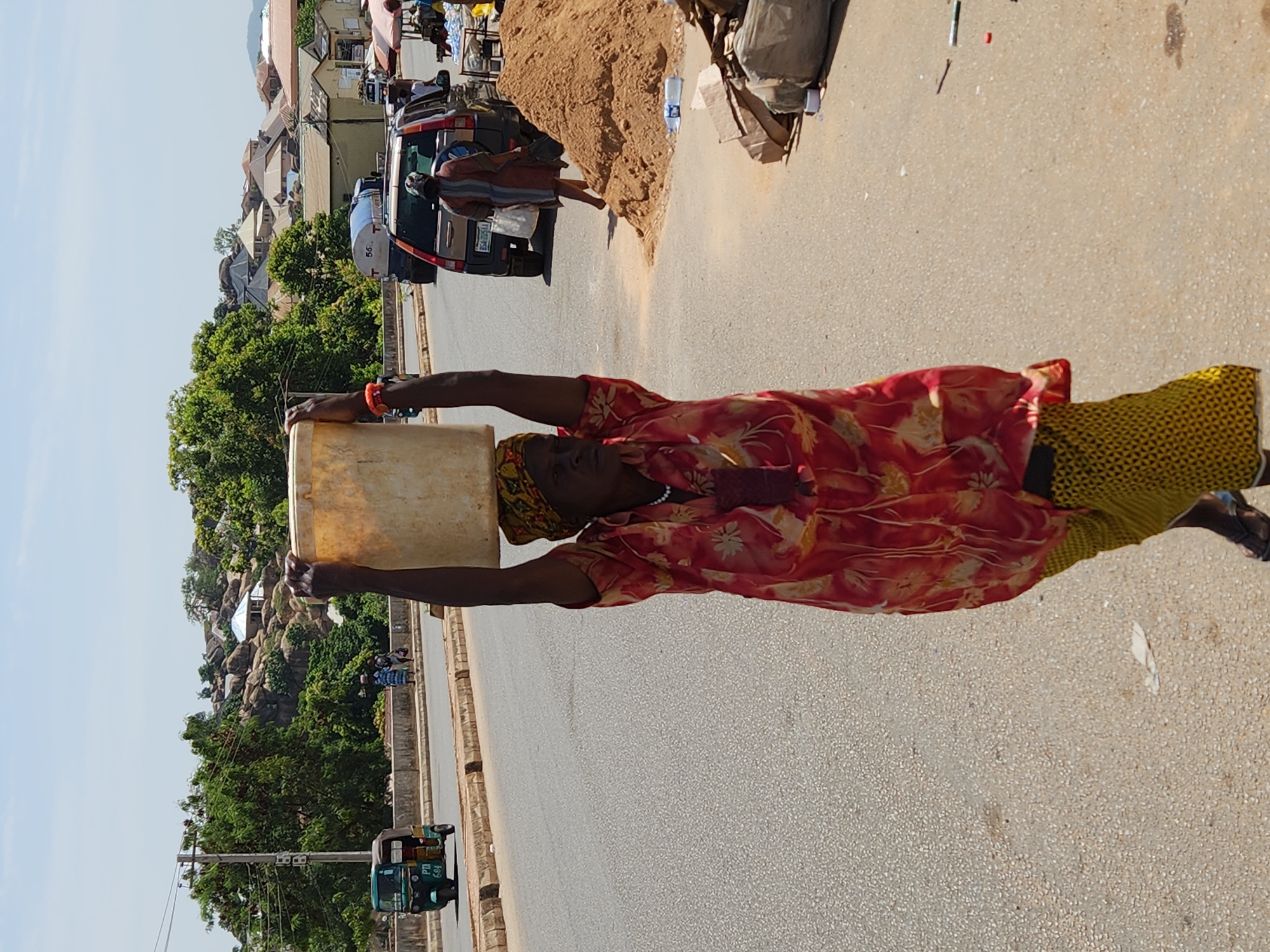 A woman is walking on a paved road in an urban area of Unknown, Unknown. She is carrying a yellow container over her head with outstretched arms. The woman is wearing a red and white patterned shirt and a yellow patterned skirt. A yellow scarf is wrapped around her head. The road is lined with buildings, trees, and parked vehicles. A pile of sand is visible next to a parked vehicle. In the background, there are other people and a green auto rickshaw.