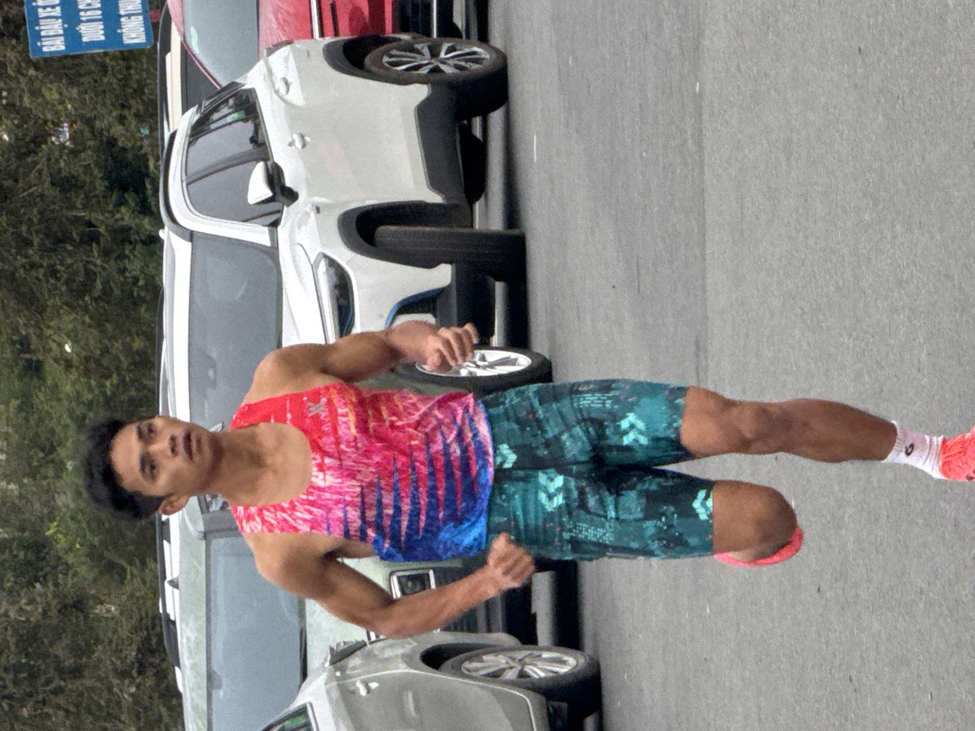 A male runner is captured mid-stride on an asphalt surface, wearing a vibrant, abstractly patterned tank top in shades of pink, blue, and red, and teal shorts with a geometric design. His right leg is extended forward, and his left leg is bent at the knee, with a pink running shoe and white sock visible. He has a focused expression and is looking forward. The background features several parked cars, including a white SUV and a red car, and a blurred backdrop of trees. A blue sign with Vietnamese text is visible on the far right. The setting appears to be an outdoor area, possibly a street or parking lot, in Da Lat, Vietnam.