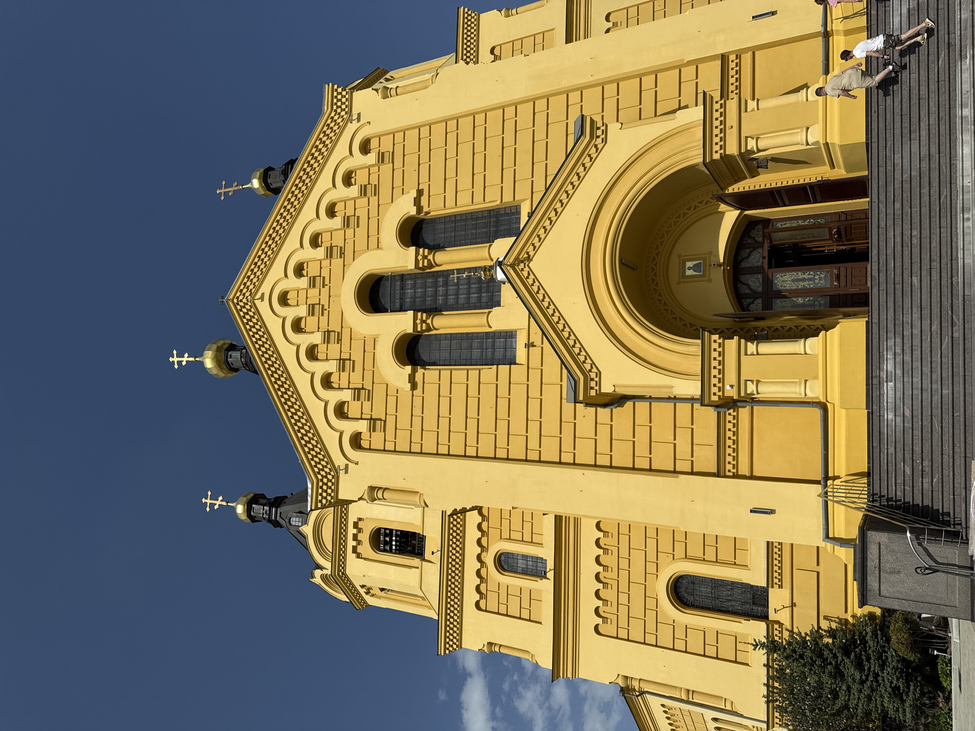 The image shows the Alexander Nevsky Cathedral in Nizhny Novgorod, Russia. The cathedral is a large, yellow building with multiple black domes topped with golden crosses. The architecture features rectangular patterns and arched windows. Two people are standing on the steps leading to the cathedral entrance. The sky is a clear, deep blue. A small tree is visible near the bottom of the building.