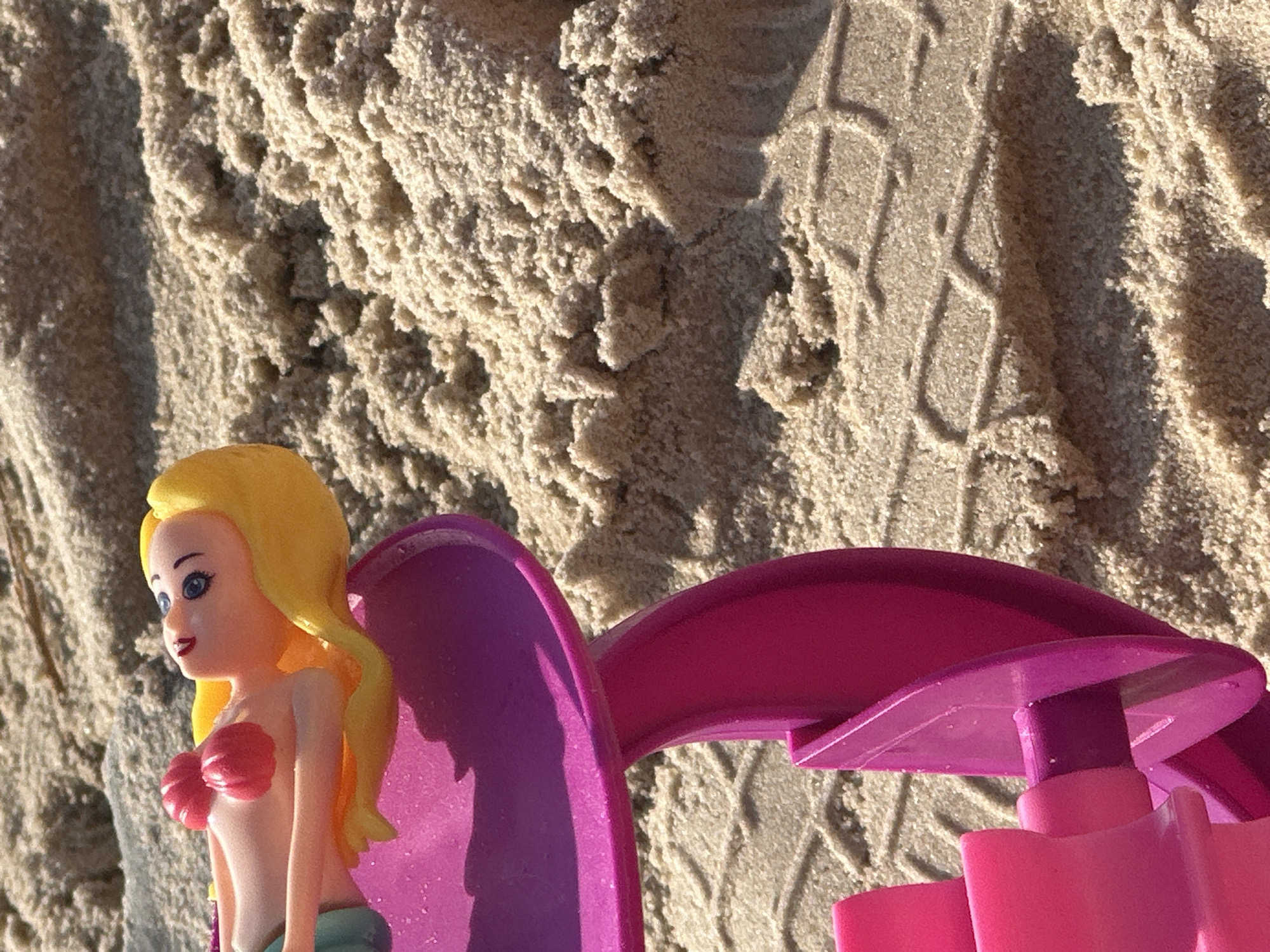 A close-up shot captures a plastic mermaid doll lying on its back on a sandy surface, possibly a beach, with a portion of a pink and purple plastic toy shovel visible in the foreground. The doll has yellow hair, blue eyes, and is wearing red seashell-shaped bikini top. The sand appears coarse and dry, with tire tracks imprinted on it. The lighting suggests it is daytime, with harsh shadows indicating bright sunlight. The overall scene suggests a playful, outdoor setting, potentially at a beach in Herzliya, Israel, given the context.