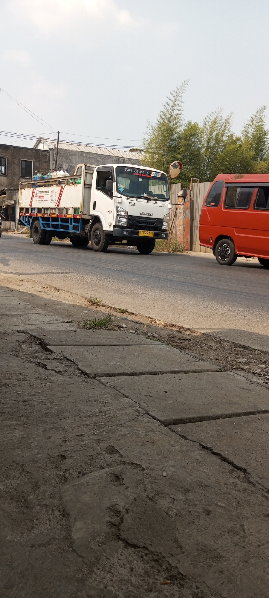 A white Isuzu Elf truck is driving on a road in Setu, Indonesia. The truck is loaded with gas cylinders and has "Gas Depot Industri" written on its side. A red van is also visible on the road. The scene appears to be in a commercial or industrial area, with buildings and trees in the background. The road is paved, and there is a sidewalk with some cracks and grass growing through it. The sky is clear and blue.
