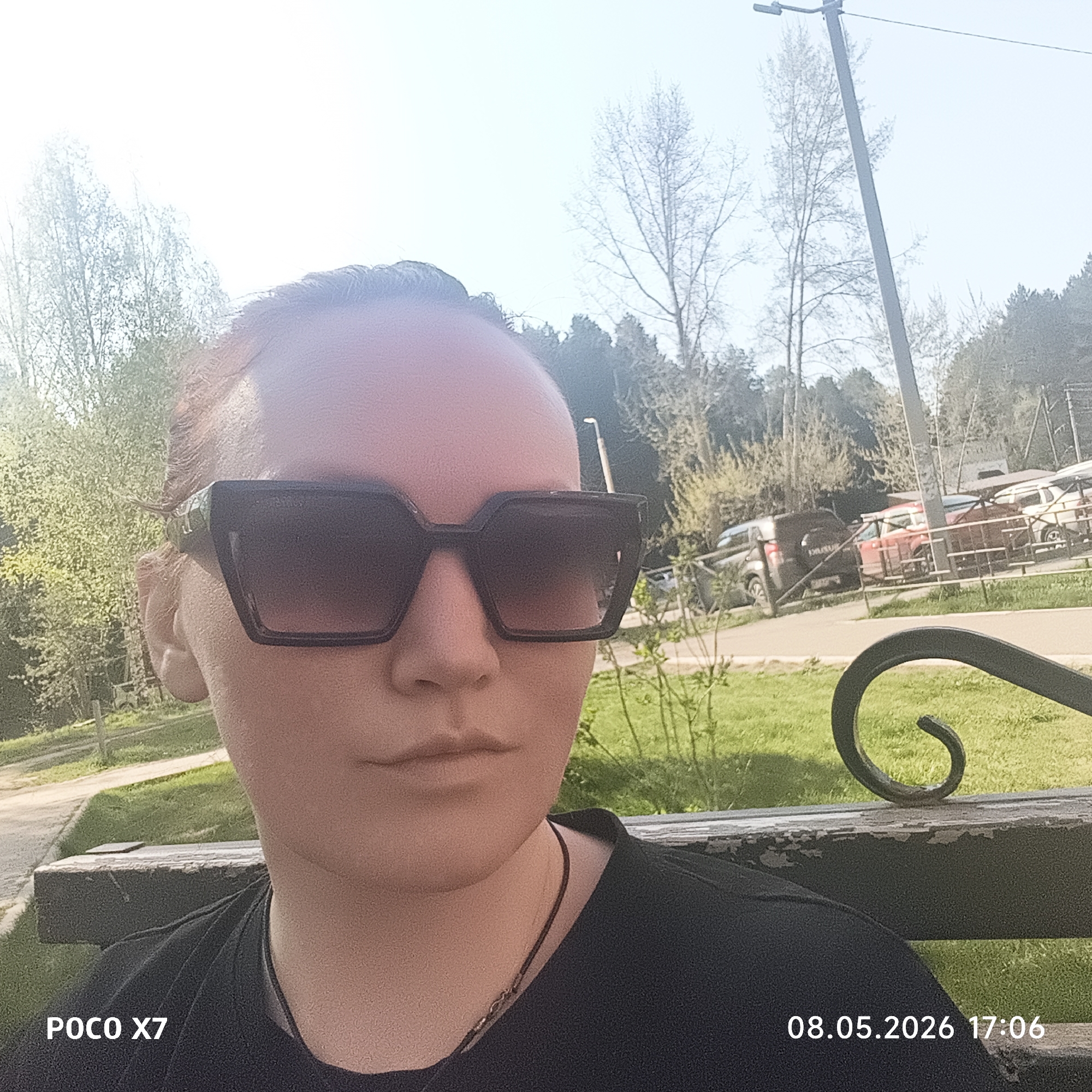 A selfie taken at 17:06 on August 5, 2026, using a POCO X7 phone. The subject is an individual wearing dark sunglasses and a dark t-shirt, sitting on a bench. Behind the subject is a grassy area with trees and some parked vehicles behind a fence. It appears to be a park or recreational area within the city of Chaikovsky, Russia, indicated by the presence of Russian license plates on the vehicles. There is a light pole visible behind the trees. The bench is painted a dark color and shows signs of wear. The subject is wearing a necklace.