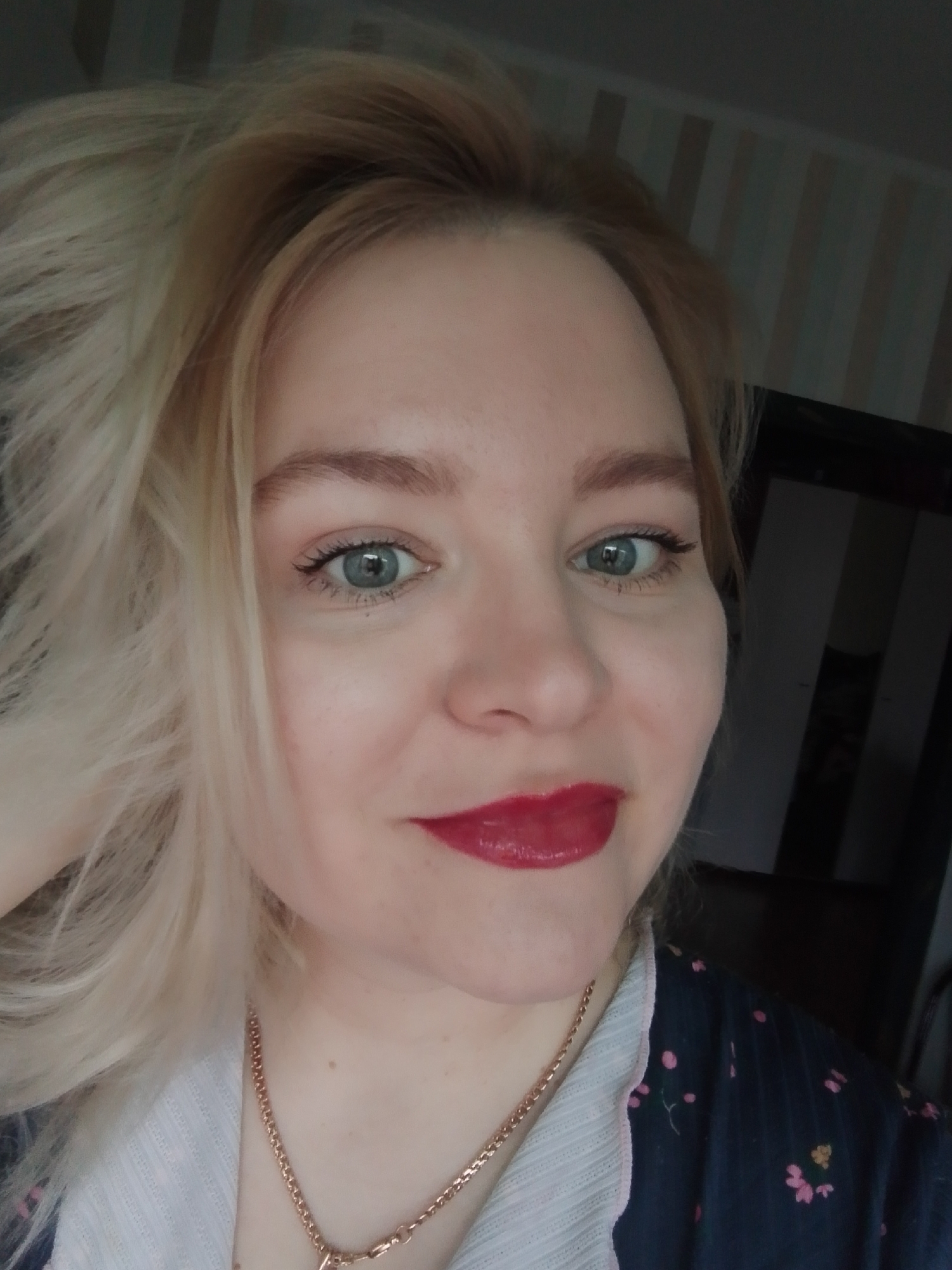 A close-up shot of a woman. She has light blonde hair, blue eyes, fair skin, and is wearing red lipstick. A gold necklace is visible around her neck. She appears to be wearing a blue floral top with a white collar. The background features striped wallpaper. A dark object, possibly a television or a doorway, is visible behind her. There is no information to associate this image with Izhevsk, Russia.