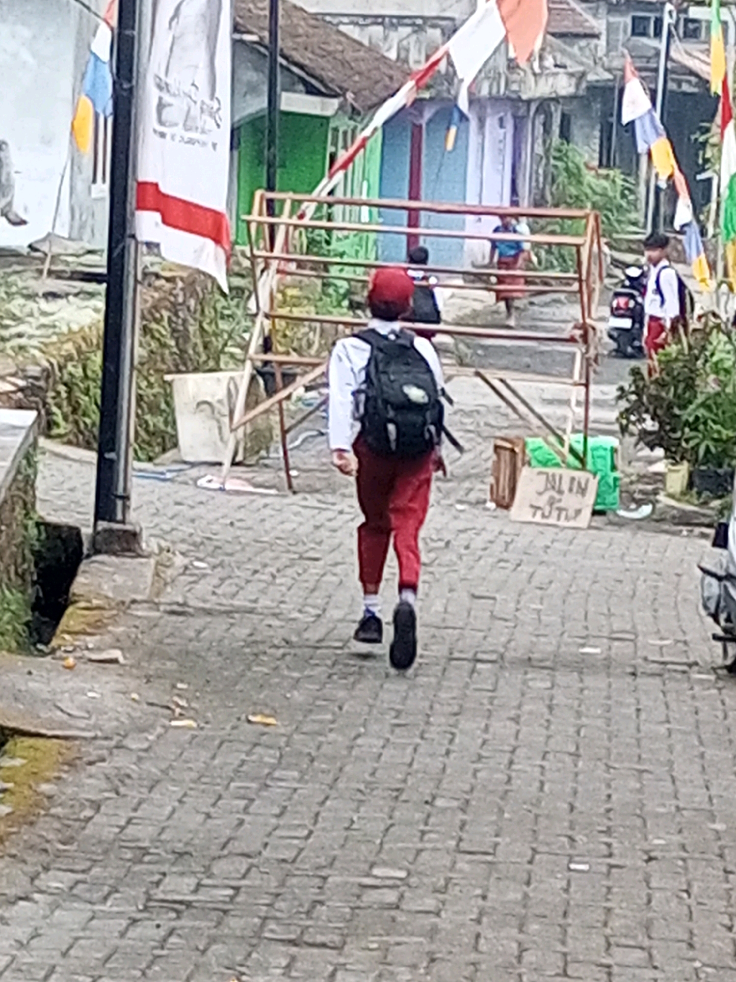 The image depicts a street scene in Andongsili, Indonesia, with a child wearing a red cap and a backpack walking away from the camera. The child is dressed in a white shirt and red pants, and is walking on a cobblestone street. In the background, there are houses with traditional Indonesian architecture, adorned with red and white flags. There is also a barrier made of metal poles and wood. A scooter is parked on the right side of the image. A sign with Indonesian writing that reads "JALAN TUTUP" (meaning "Road Closed") is visible near the barrier. The overall environment suggests a village or a residential area within the city, with a festive atmosphere indicated by the flags.