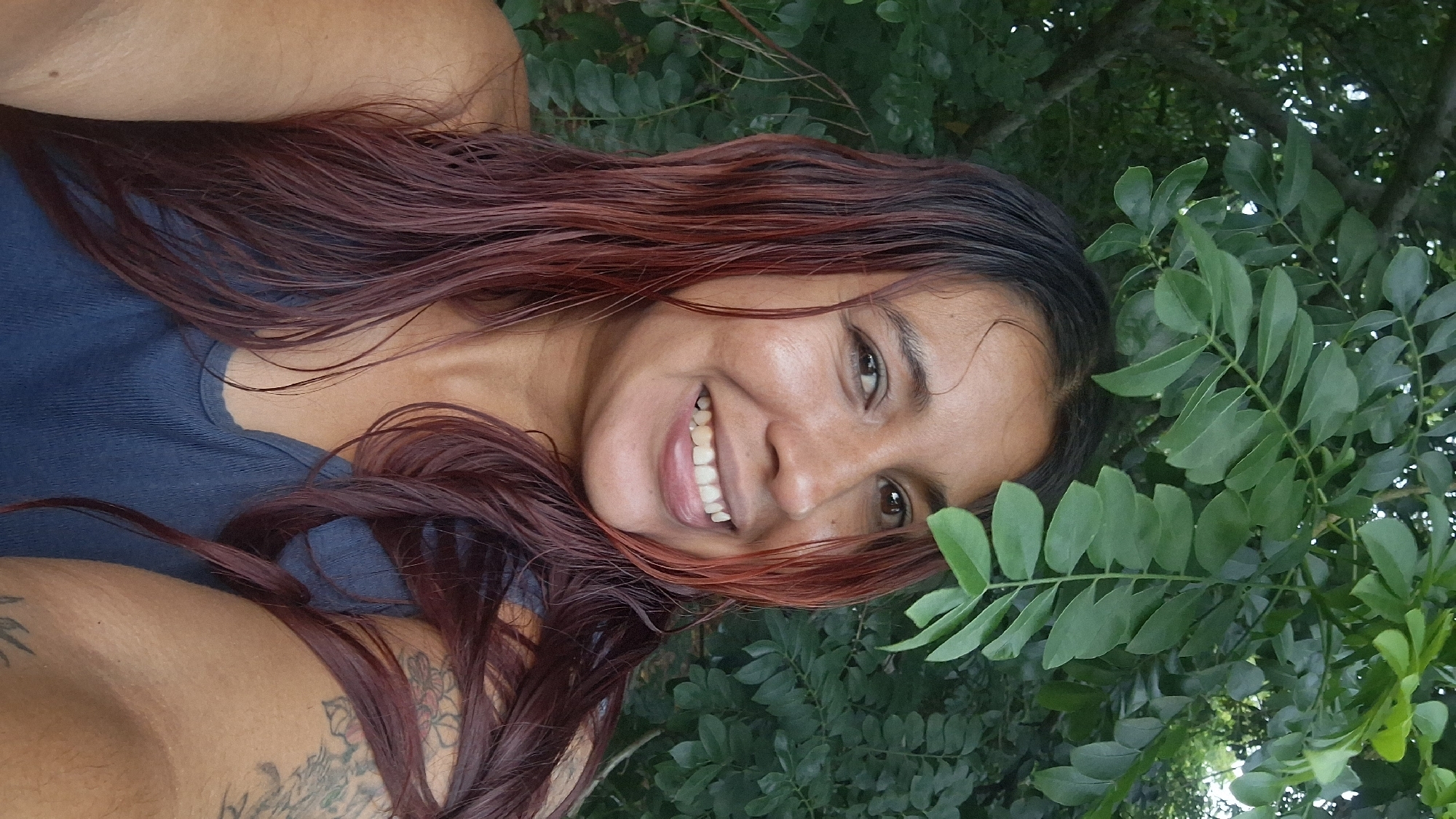 In Vallecito, Venezuela, a woman with long, dark red hair is captured in a close-up portrait outdoors. She is wearing a blue tank top and has a tattoo on her right arm. She is smiling broadly, with her teeth showing. Green leaves from a tree are visible in the foreground, partially obscuring the top of her head and framing her face. The background is filled with lush green foliage. The lighting suggests it is daytime.