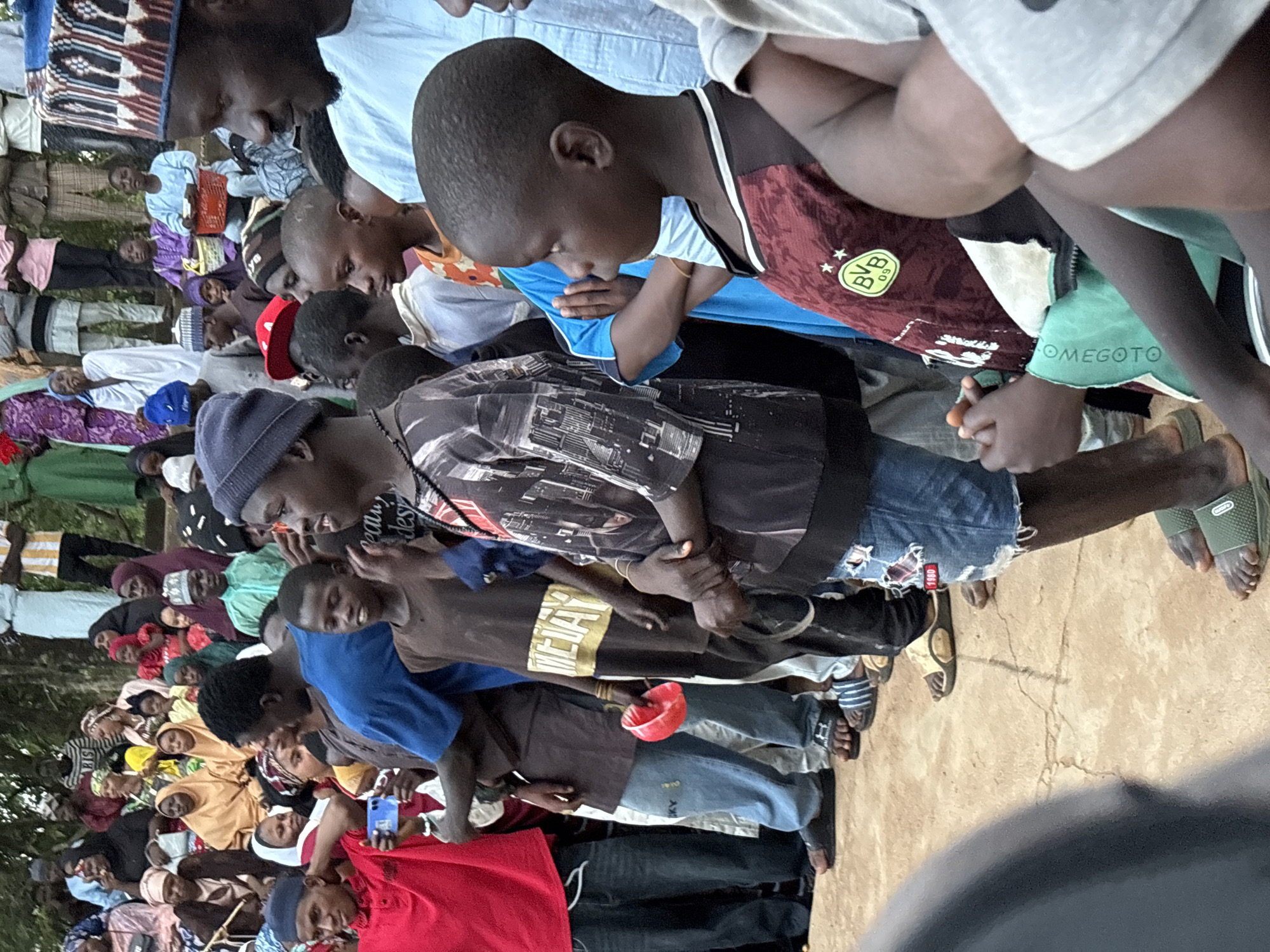 A group of people are gathered outdoors on a dusty, cracked earth surface. The individuals are of various ages, with many children and young adults visible. They are dressed in a mix of traditional and modern clothing, including headscarves, long shirts, t-shirts, and denim shorts. Some people are holding personal items such as a red plastic bowl and a smartphone. The atmosphere appears to be social, with people standing and facing in different directions, suggesting a gathering or event. The background shows hints of lush green foliage and possibly buildings, suggesting a semi-urban or suburban environment. The lighting indicates it is daytime, with no strong shadows suggesting direct sunlight. The overall scene depicts a community event or gathering in Jos, Nigeria.