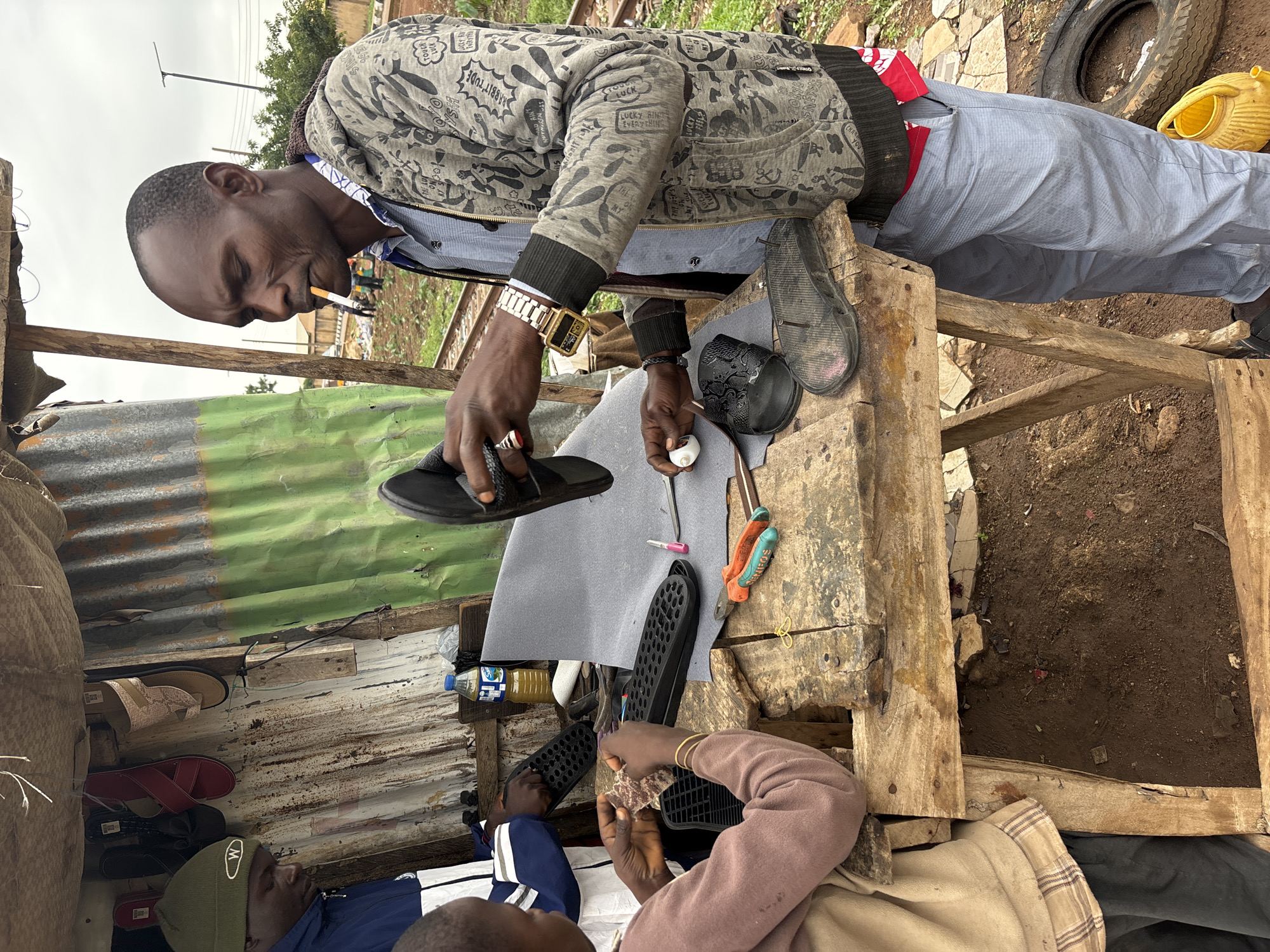 In Barakin Lerawi, Nigeria, an outdoor workshop is set up on a wooden table, with a backdrop of corrugated metal and green sheeting. A man, with a cigarette in his mouth, is working on a sandal. He is wearing a patterned grey hooded jacket over a blue shirt and grey trousers, and a golden watch on his left wrist. Next to him, a child in a light brown jacket is also working on a sandal. Various tools, including orange and green pliers, scissors, and a white circular object, are scattered on the table. Several finished and unfinished sandals are also present, along with a bottle of liquid and a roll of grey material. In the background, a pair of train tracks and some distant structures can be seen, suggesting the workshop is located near a railway line. The ground is covered in dirt and debris, with a deflated tire and a yellow watering can visible.