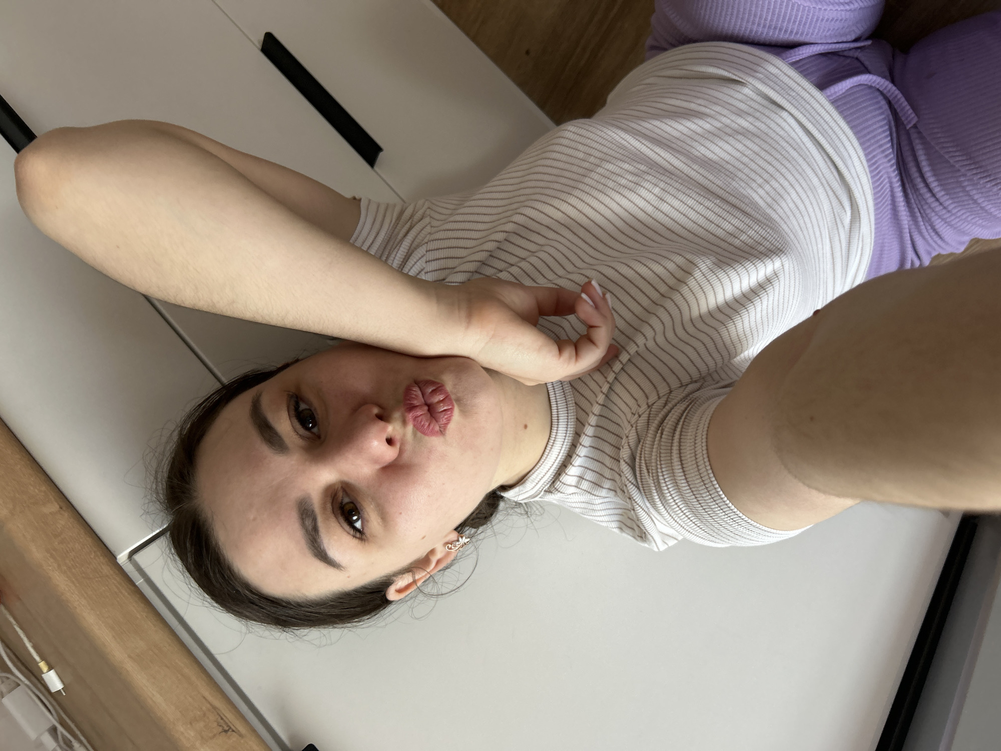 A young woman is lying on a white surface, possibly a dresser, wearing a white striped shirt and purple pants. She is facing the camera with her lips pursed in a kissing gesture. Her right arm is raised near her head, and her left arm is extended towards the camera, presumably holding it for a selfie. The environment appears to be indoors, with wooden and white furniture visible. There is no clear indication of this image's location as Chaikovsky, Russia.