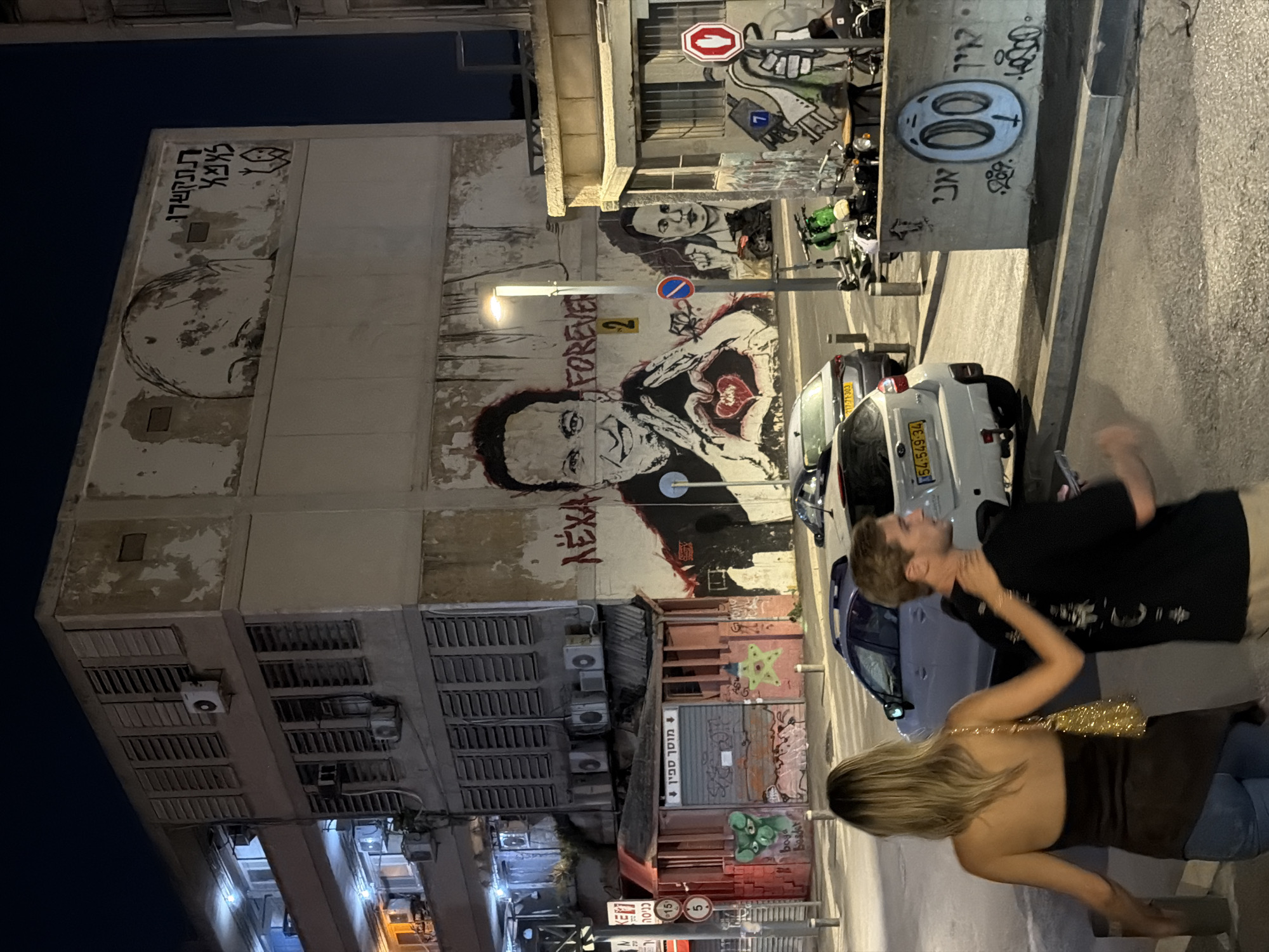 The image captures an urban street scene at night in Tel-Aviv, Israel.
The dominant feature is a multi-story, somewhat deteriorated concrete building with peeling paint, exposed air conditioning units, and horizontal louvered window coverings. A large mural on its side depicts a smiling man with dark hair forming a heart shape with his hands, with a red heart in the center. Text next to him reads "NEXA" and "FOREVER 2". To the right of this, a smaller mural shows a woman's face. Near the top of the building, a sketch of a bald head is accompanied by Hebrew text "תתקשרו למצוא" (Titkashru Limtzo - Call to find/locate) and a "call me" hand gesture symbol.
At street level on the left, a small, red-painted structure with a closed metal shutter features graffiti, including a green cartoonish face and a yellow star. Hebrew text on this structure identifies it as "מוסך ספין" (Mosakh Spin - Spin Garage) with an "entrance" sign.
On the right side of the street, a light-colored concrete low wall displays graffiti, including the Hebrew word "אני" (Ani - I am) next to a stylized circular face. A streetlight illuminates this area. Numerous scooters and at least one motorcycle are parked on the sidewalk. A red stop sign with a white hand icon is visible.
Two cars are parked on the street: a white Subaru SUV (Israeli license plate 54-549-34) in the mid-ground and a silver sedan (Israeli license plate 217-71-303) behind it.
In the foreground, two individuals are walking away from the viewer. A woman with blonde hair, wearing a dark, asymmetric halter-neck top and blue jeans, carries a golden, sparkly handbag. Her left hand is placed on the back of a man walking beside her. The man has light brown hair, wears a black t-shirt with a white graphic design on the back, and light-colored pants, holding a mobile phone in his right hand. The street is paved with visible curbs and sidewalks.