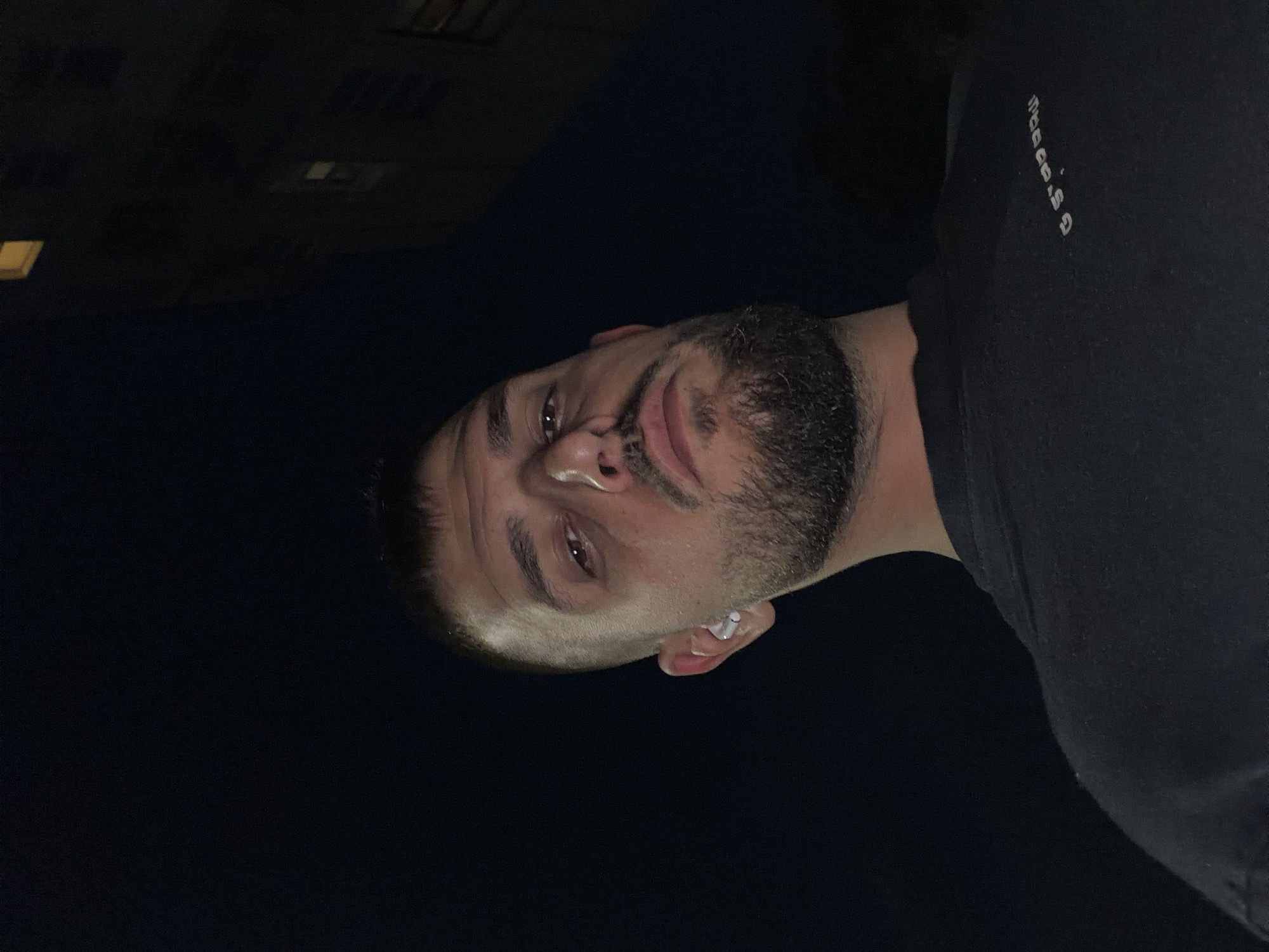 A medium close-up, taken at night in Sofia, Bulgaria, captures a man's face and upper torso. The image is oriented vertically, with the man's head tilted to the right within the frame. He has short dark hair and a neatly trimmed beard, and is looking directly at the camera with a neutral to slightly pensive or smirking expression. A white earbud is visible in his right ear. He is wearing a dark, possibly black, t-shirt, which has small, stylized white text on the right side of the chest that reads "thoooo.G".
The setting is outdoors at night, with the background being largely dark and indistinct. Faint, rectangular lights, likely from windows of a building, are visible in the upper left background, suggesting an urban environment. The man's face is illuminated, possibly by a flash or direct light source, creating highlights on his skin and eyes, while the rest of the scene remains dark. The overall mood is intimate and casual, with the darkness emphasizing the subject.