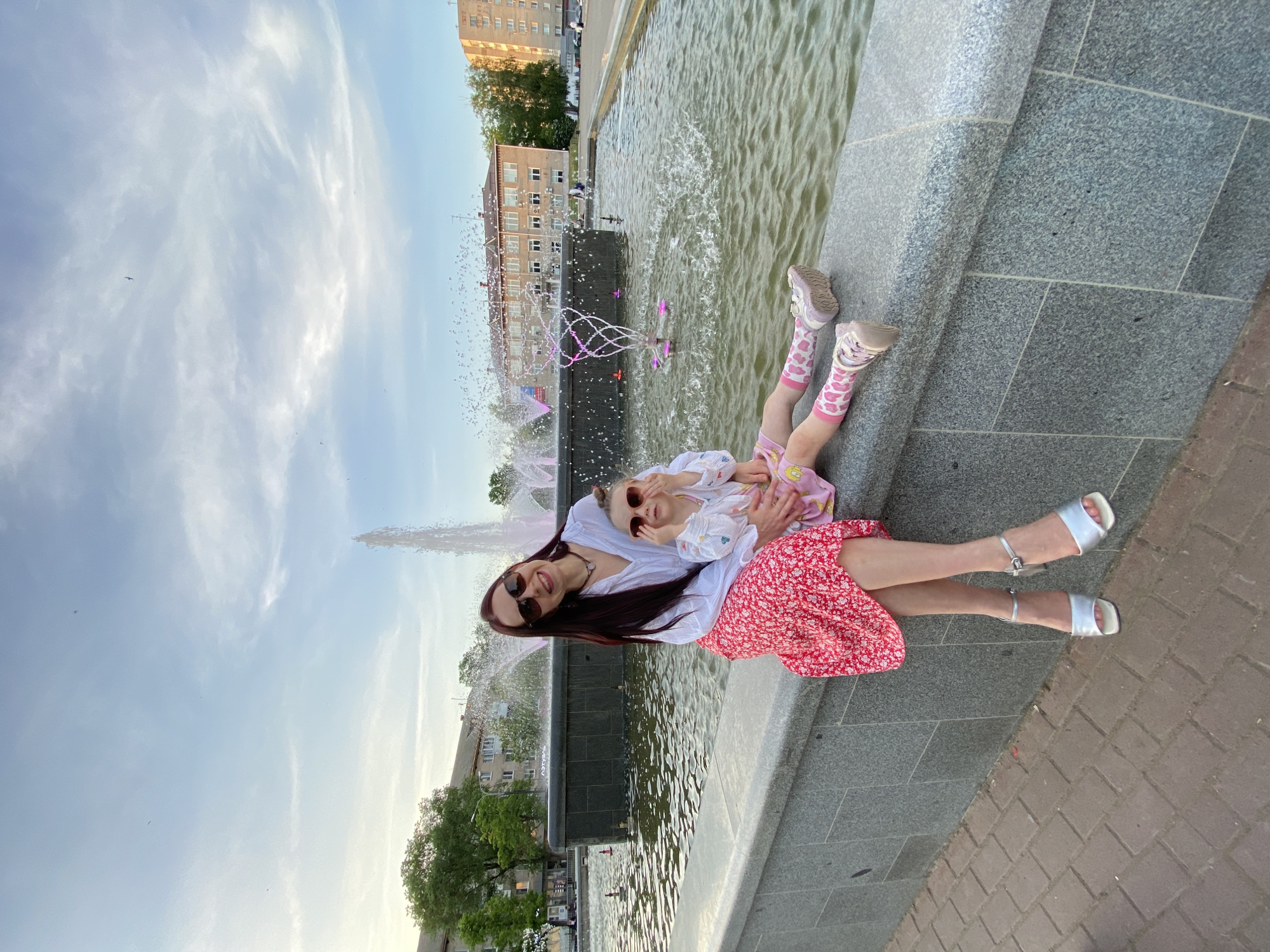 A medium shot captures a woman and a child posing beside a fountain. The woman, with long dark hair and sunglasses, wears a white blouse and a red floral skirt, along with silver sandals. She is seated on the edge of the fountain, holding a young child in her lap. The child, also wearing sunglasses, is dressed in a white top, a pink dress, pink socks with hearts, and pink shoes.
The fountain has a large vertical water spout as well as smaller water features tinged with purple light. Behind the fountain is a multi-story brick building. The sky is light blue with some wispy clouds. The scene is located in the city of Chaikovsky, Russia. The location is evidenced by information visible in the image.