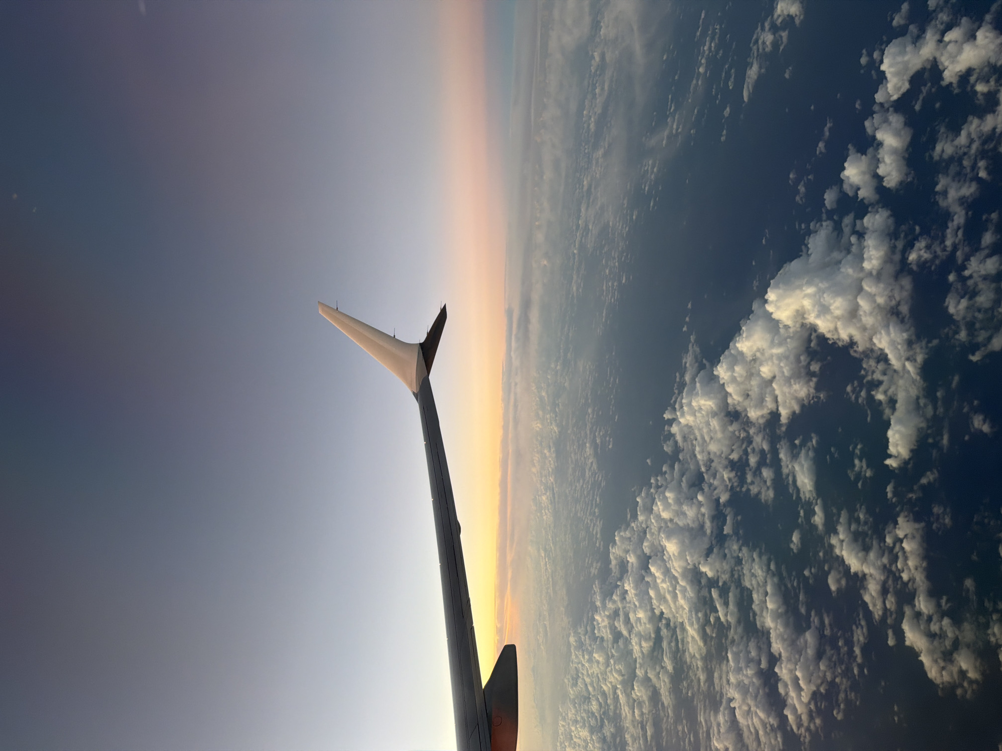 The image shows a view from an airplane window. The wing of the plane is visible in the foreground, extending diagonally from the bottom to the left. The sky above is a gradient of colors, transitioning from a soft blue to a warm orange-yellow horizon. Below the horizon, the landscape is covered in clouds, creating a textured and varied pattern of white and dark blue. The angle of the image suggests the plane is in flight.