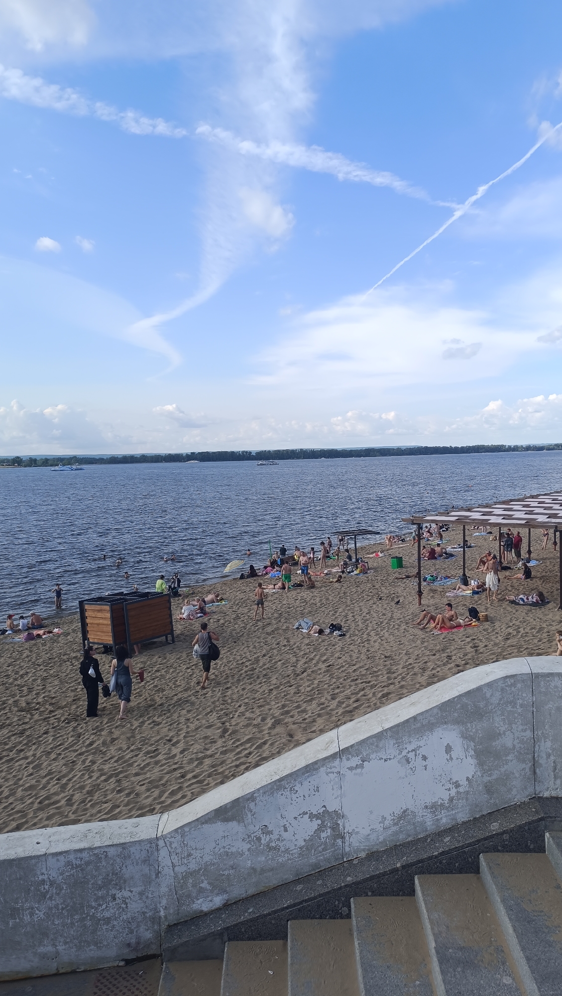 The image depicts a crowded beach on a sunny day. The beach is sandy, with many people sunbathing, swimming in the water, and walking around. There are several umbrellas and canopies providing shade. In the background, a wide river stretches to the horizon, with trees lining the opposite bank. Several boats are visible on the water. In the foreground, a concrete structure with stairs leads down to the beach. The sky is blue with scattered white clouds and contrails from airplanes. This scene is characteristic of a public beach in Novokuybyshevsk, Russia, likely along the Samara River or a similar large body of water.