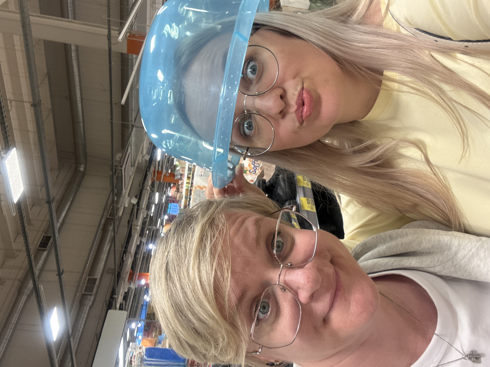 In an indoor supermarket, two individuals pose for a selfie. The person on the left, with short blonde hair and glasses, is looking at the camera with a neutral expression. The person on the right, with long blonde hair, is wearing a blue plastic dome-like object on their head and is puckering their lips towards the camera. Both individuals appear to be casually dressed. The background shows typical supermarket aisles with shelves stocked with various products, and fluorescent lighting illuminates the scene. The overall atmosphere is lighthearted and playful.