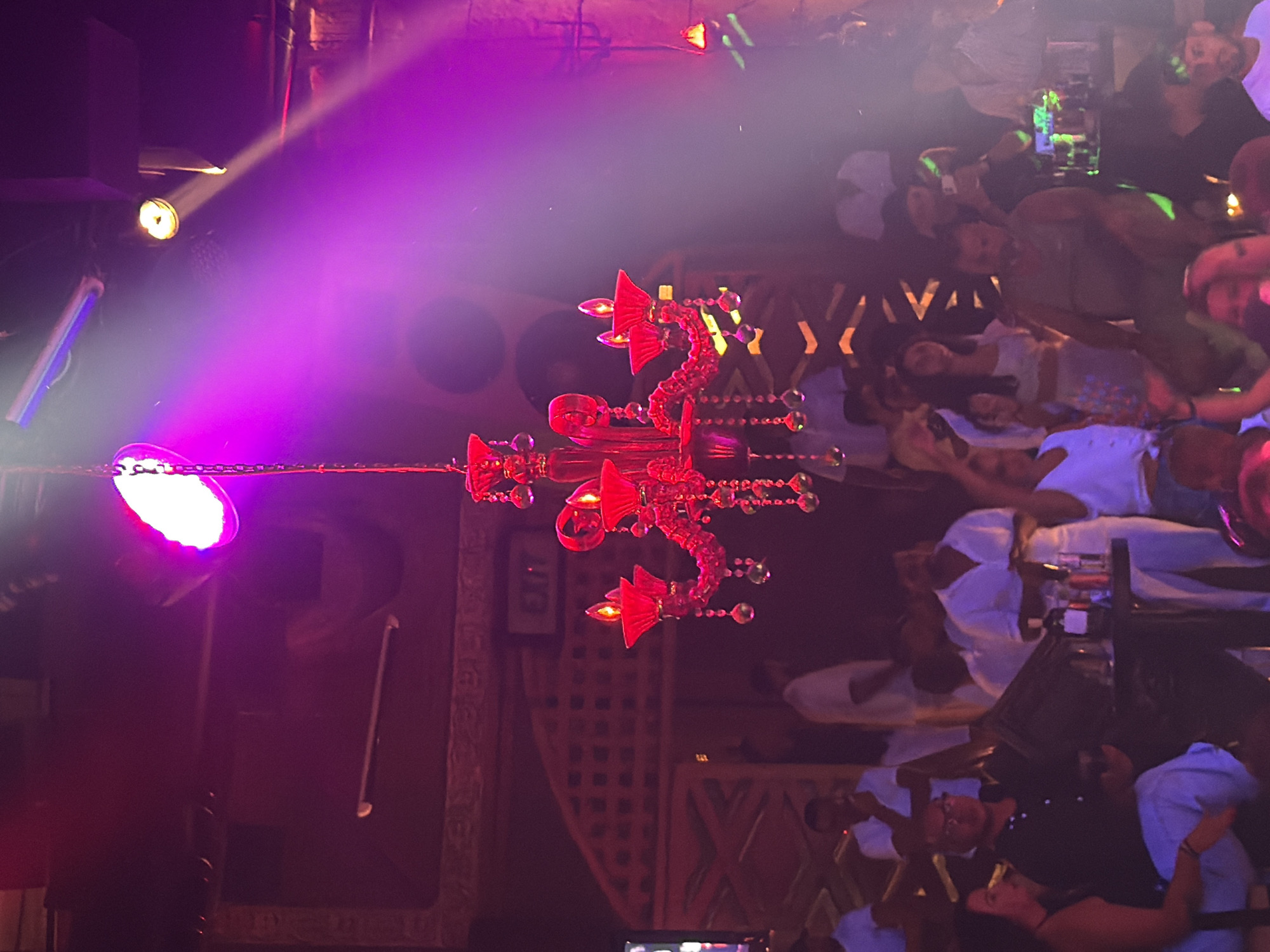 The image depicts an indoor nightclub scene with a vibrant, dimly lit atmosphere, predominantly characterized by purple and pink lighting. A prominent bright pink light fixture is visible at the top center of the frame, casting beams of light across the room. In the foreground, a large, red, elaborate chandelier hangs, adorned with sparkling crystal-like ornaments and red candle-shaped lights.
The nightclub appears to be moderately crowded with individuals dressed in casual attire. Some are seated, while others are standing and socializing. A table with drinks and bottles is visible on the right side of the image. In the background, a wall with a textured, decorative pattern can be seen, along with an "EXIT" sign illuminated in red. The overall ambiance suggests a lively evening entertainment venue, likely in a popular tourist destination such as Sharm El Sheikh, Egypt, known for its vibrant nightlife.