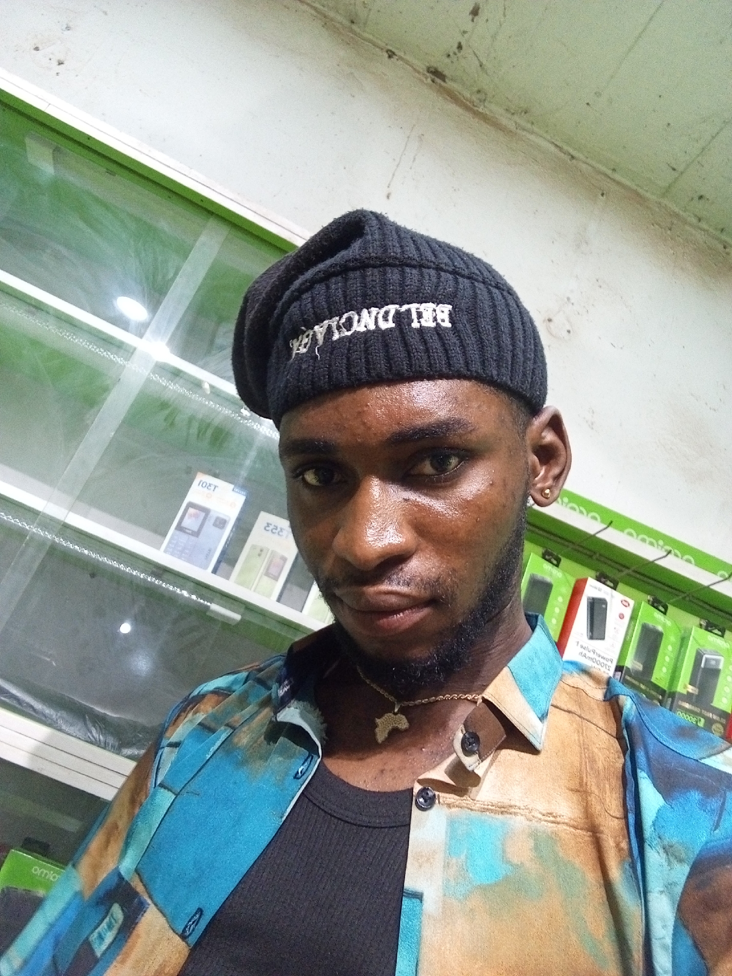A medium shot, taken from a low angle, shows a Black male in Lokoja, Nigeria, standing in front of a display of mobile phones. The man is wearing a black knitted beanie with white lettering, a black undershirt, and a brightly colored patterned shirt with blue, tan, and brown hues. He is also wearing a gold chain with a pendant in the shape of Africa. The background consists of shelves filled with boxed mobile phones, illuminated by fluorescent lighting. The overall lighting is dim, with reflections visible on the glass display cases. The image is a selfie, with the camera positioned slightly above and to the left of the subject.