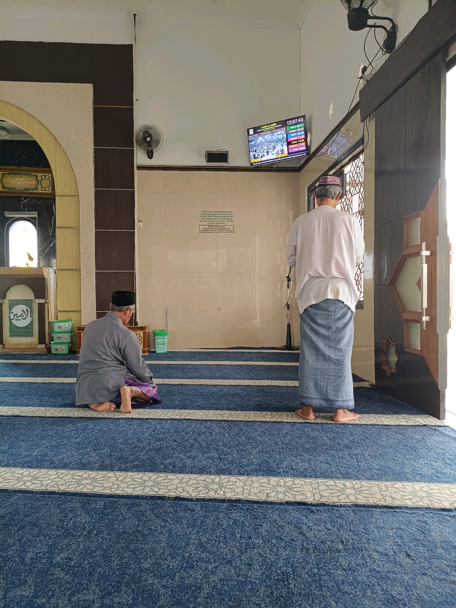 The image depicts the interior of a mosque in Maguwoharjo, Indonesia, with two men engaged in prayer. The mosque features blue carpet with white patterned stripes as the floor covering. On the left side of the image, one man is kneeling on the carpet, facing away from the camera, wearing a grey thobe and a patterned sarong. To the right, another man stands, also facing away, dressed in a white thobe and a blue patterned sarong. Behind him, a television screen displays prayer times and images of Mecca. The wall behind the standing man is tiled, and an air circulator is mounted on the wall to the left. An arched doorway with patterned glass is visible on the right side of the frame. The lighting suggests daylight filtering into the mosque.