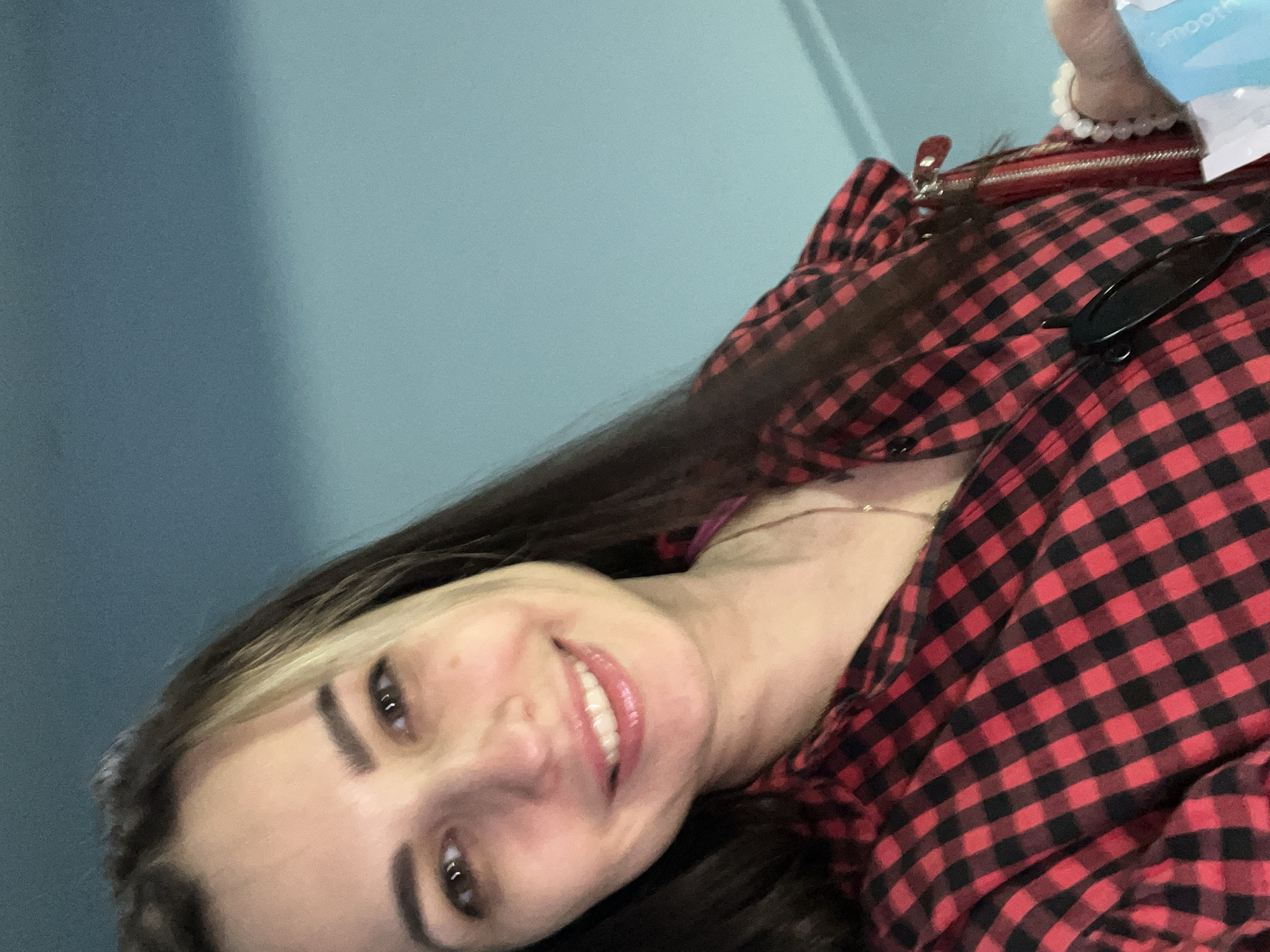 A close-up, slightly rotated portrait features a smiling woman with long, dark hair, some strands of which are lighter near her face. She is looking directly at the camera with a bright, open-mouthed smile, revealing her teeth. She is wearing a red and black buffalo plaid button-down shirt. Black sunglasses are hooked onto the collar of her shirt.
In the upper right corner, a red purse or small bag with a zipper is partially visible. Her right wrist, wearing a white beaded bracelet, is also seen, holding a light blue bottle or container with the partially visible word "smooth" printed on it. A thin necklace is faintly visible around her neck.
The background consists of a plain wall or surface, primarily light blue, with a darker blue section on the left side of the image, suggesting an indoor setting. The overall impression is a casual, cheerful moment captured in a selfie.