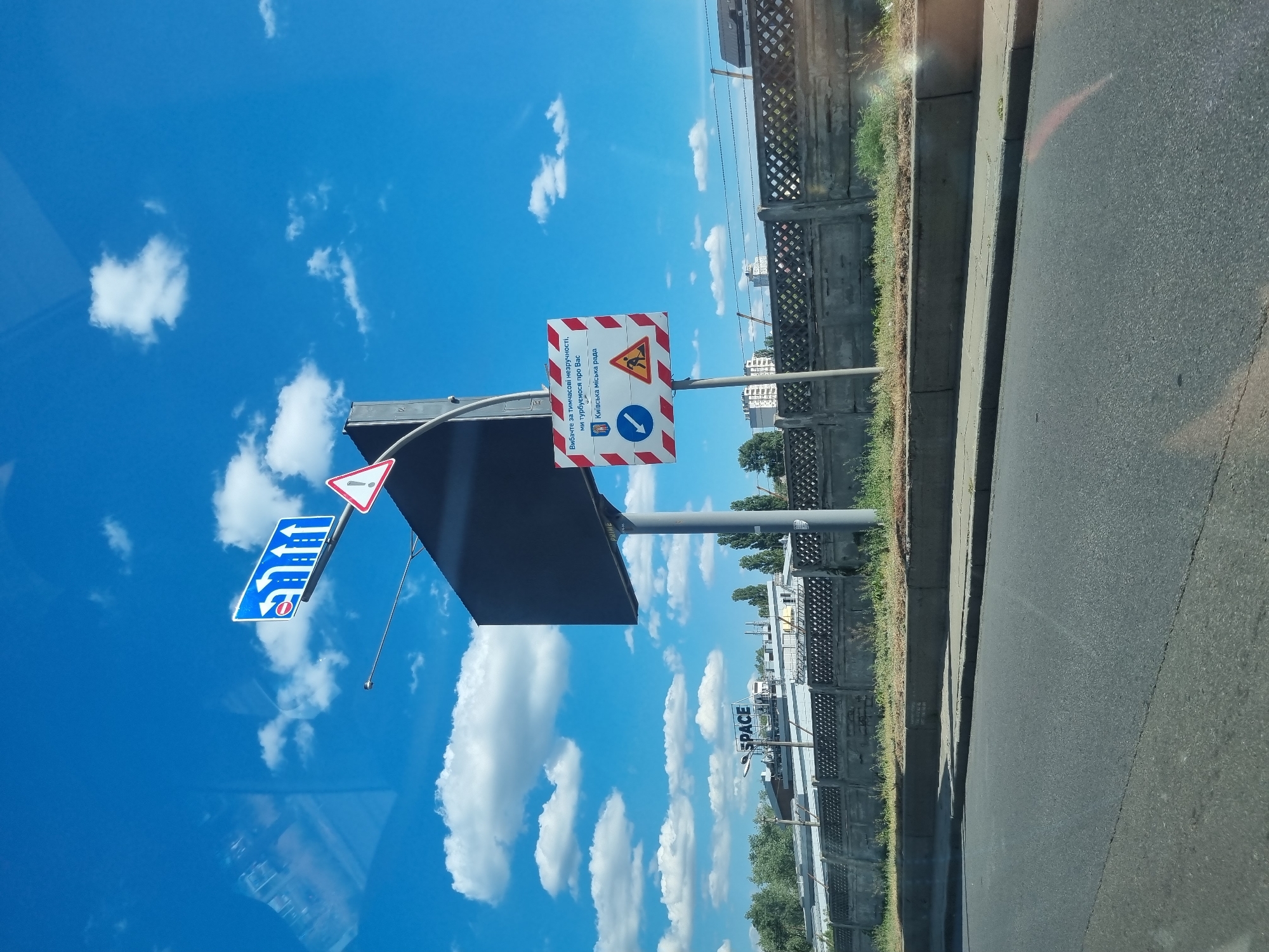 The image shows a roadside scene in Kyiv, Ukraine, under a bright blue sky with scattered white clouds. A tall pole supports a large black billboard on one side and two traffic signs on the other. The traffic signs are a combination of informational and warning signs. One sign, with red and white diagonal stripes, contains text in Ukrainian, an emblem of the city of Kyiv, a blue circle with a white arrow pointing left and a red diagonal bar, and a yellow triangular warning sign with a black silhouette of a person working. The other sign is a blue rectangular sign with white arrows indicating lanes and a red circle with a diagonal bar over a white arrow, signifying no entry.
In the background, there is a concrete fence with a diamond pattern, a low-lying building with the word "SPACE" visible on its rooftop, and beyond that, some trees and apartment buildings. The foreground shows a paved road and a curb. The overall impression is an urban environment with road infrastructure and advertising.