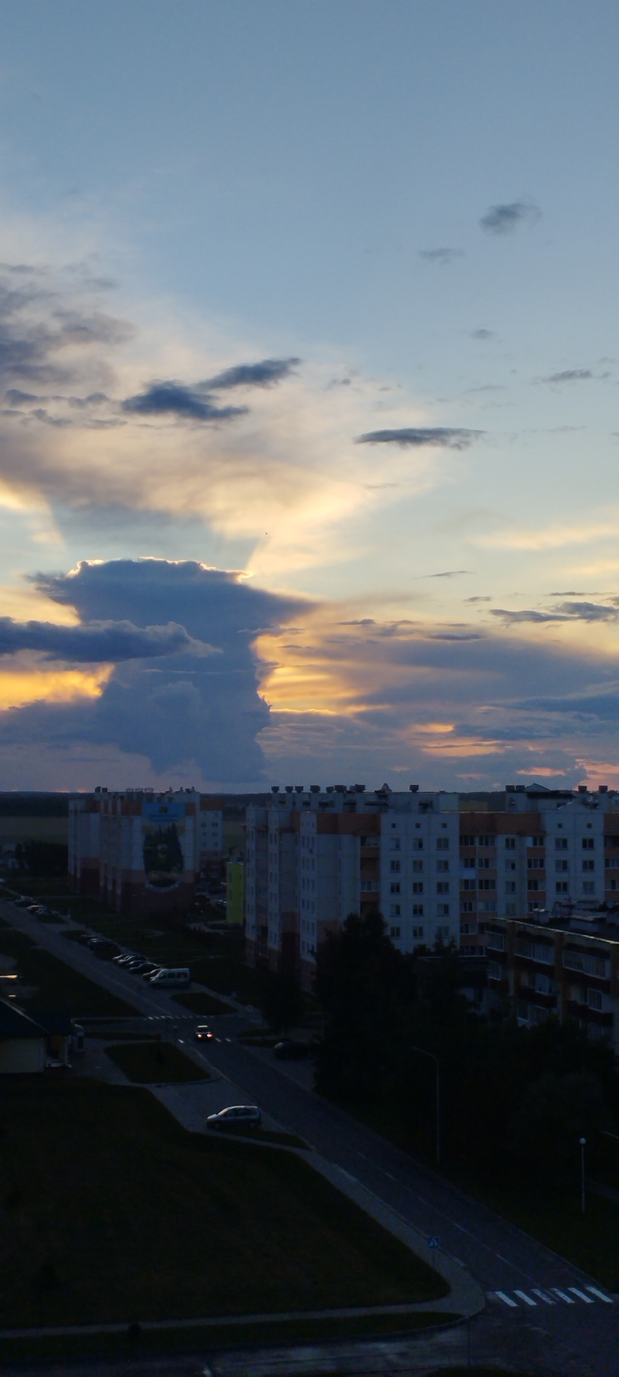 This is a vertical, full-shot photograph taken at dusk in the city of Skidzieĺ, Belarus. The image depicts a broad view of apartment buildings and a street scene, with a dramatic sky filling the upper portion of the frame. The sky is filled with colorful clouds, transitioning from a light blue at the top to shades of orange, yellow, and gray closer to the horizon. A large, dark cloud formation dominates the center-left, with shafts of light breaking through it.
In the middle ground, a row of multi-story apartment buildings stretches across the frame. These buildings appear to be residential, with numerous windows visible. Some of the buildings have light-colored facades with orange trim. A mural is visible on the side of one building.
In the foreground and lower middle ground, a paved road runs through the scene, lined with parked cars. A few vehicles are in motion on the road, including one with its headlights on. Streetlights are present, illuminating parts of the road and surrounding grass areas. The overall impression is that of a typical urban evening scene.