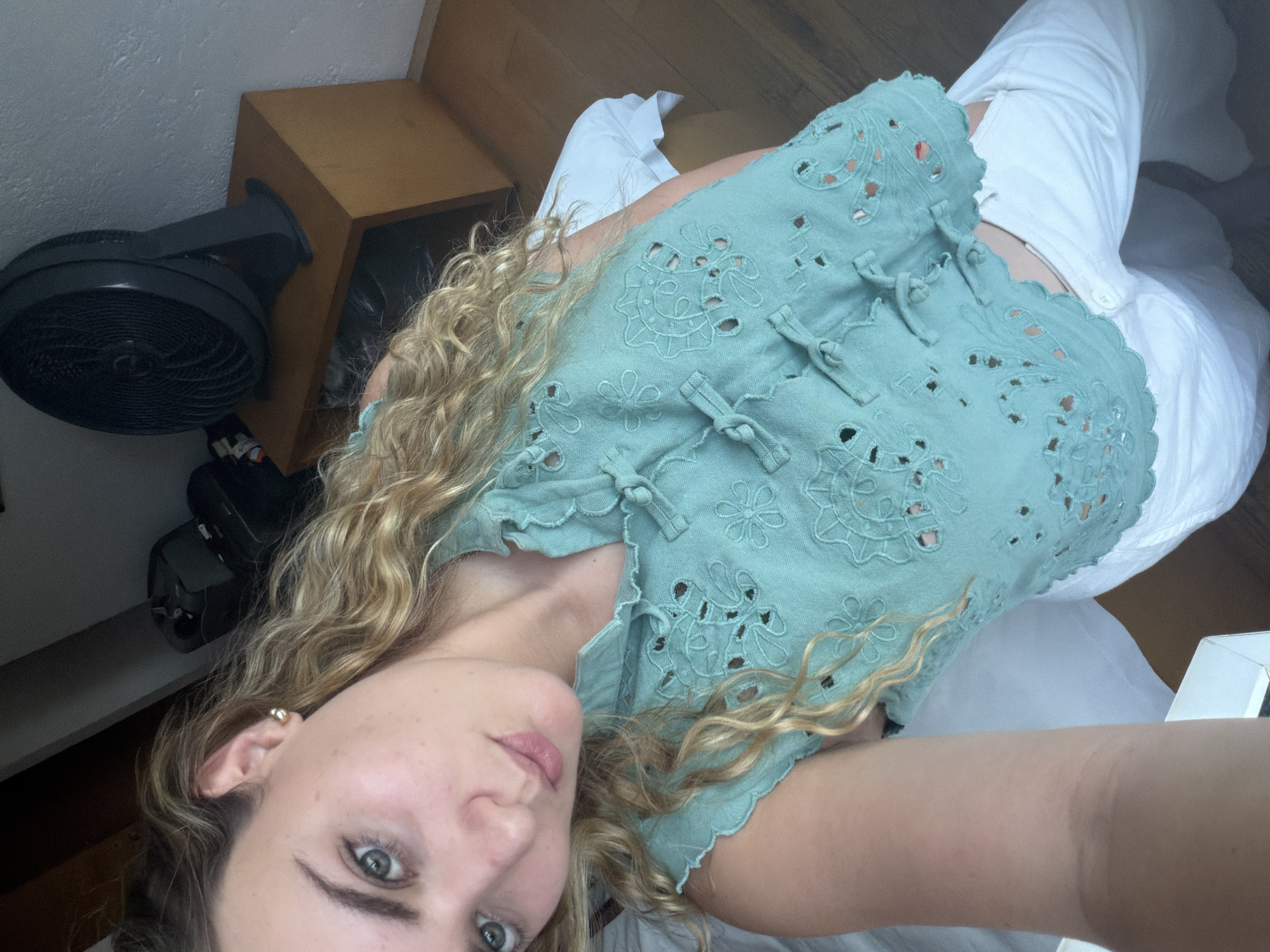 The image, oriented horizontally, captures a young woman in an indoor setting. She has long, wavy blonde hair that cascades around her shoulders and blue eyes, looking directly towards the viewer with a neutral and calm expression. She wears a light teal or mint green top featuring intricate eyelet embroidery and several decorative frog closures down the front. The fabric of her top has a textured appearance. Her lower body is clad in white trousers or pants, part of which is visible, showing a white button detail.
In the background to her left, a black floor fan is positioned next to a light brown wooden cabinet or nightstand. To her right and behind her, white bedding or fabric is visible, along with a small, clear, possibly acrylic object. The floor appears to be constructed from wooden planks. She wears a small gold stud earring in her visible ear. The lighting suggests an indoor daytime setting with ambient light. There is no visible text in the image. The provided location context for this scene is San Bernardino Tlaxcalancingo, Mexico.