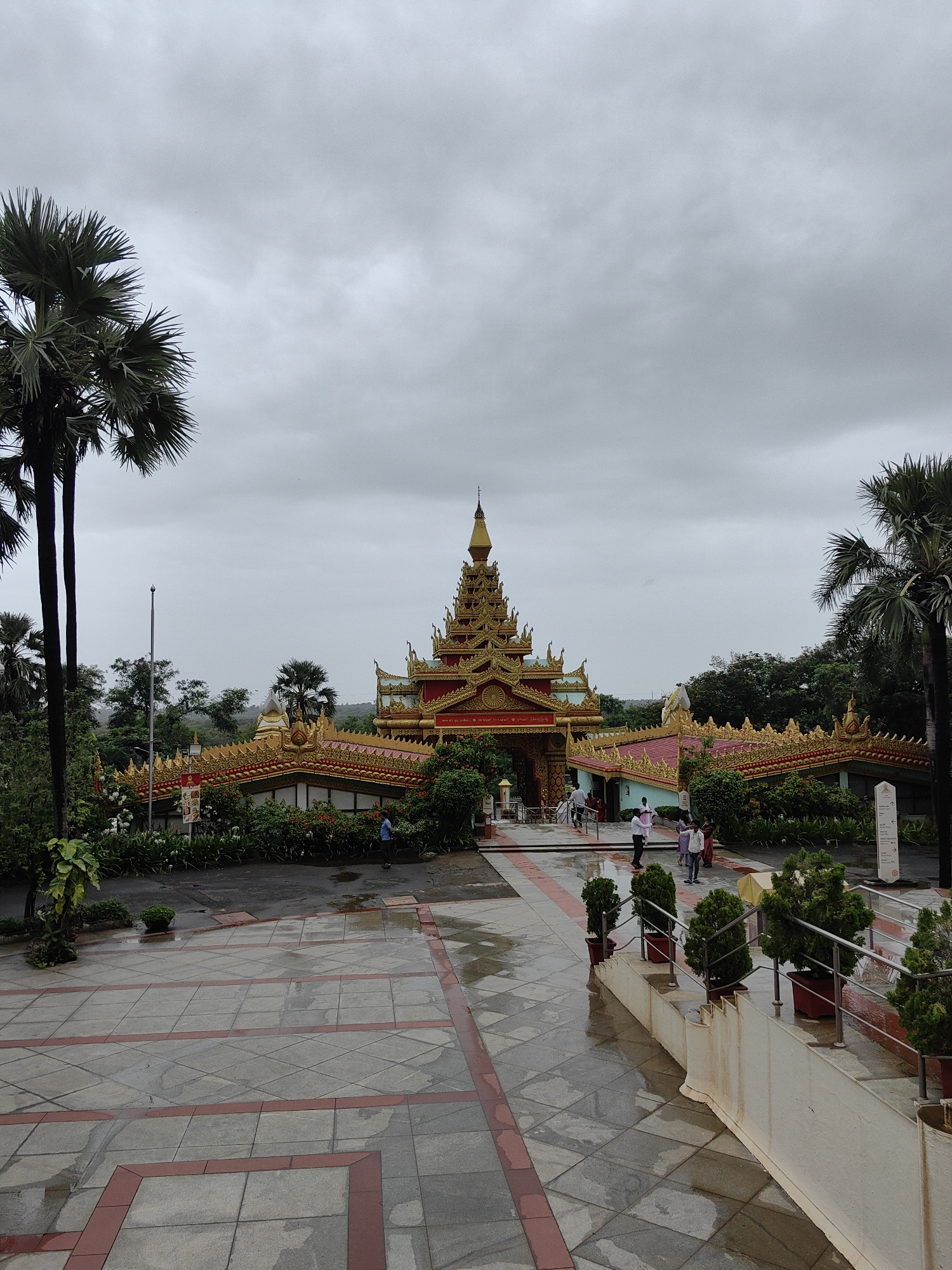The image captures the entrance of the Global Vipassana Pagoda in Mumbai, India, on an overcast day. The imposing structure features a central golden dome topped with a spire, surrounded by elaborate golden and red tiered roofs. The architecture is distinctly Buddhist, with intricate decorative patterns and pointed finials.
In the foreground, a paved courtyard with a pattern of grey tiles and red borders reflects the dampness from recent rain, suggesting a humid environment. Several potted evergreen shrubs are arranged along a white retaining wall to the right, with metal railings guiding a gentle slope.
To the left of the pagoda, tall palm trees with their fronds reaching towards the sky frame the scene. Greenery and manicured bushes line the pathway leading to the entrance, with red and pink flowers adding touches of color.
Several people are visible in the middle ground, walking towards or away from the pagoda entrance. A man in a blue shirt is seen walking on the wet pavement to the far right. Others, dressed in light-colored clothing, are near the entrance area.
The background is filled with lush green trees, indicating a location with abundant vegetation, characteristic of parts of Mumbai. The sky is heavily clouded, creating a diffused light and a muted atmosphere. The overall scene conveys a sense of a spiritual and serene place, despite the overcast weather.