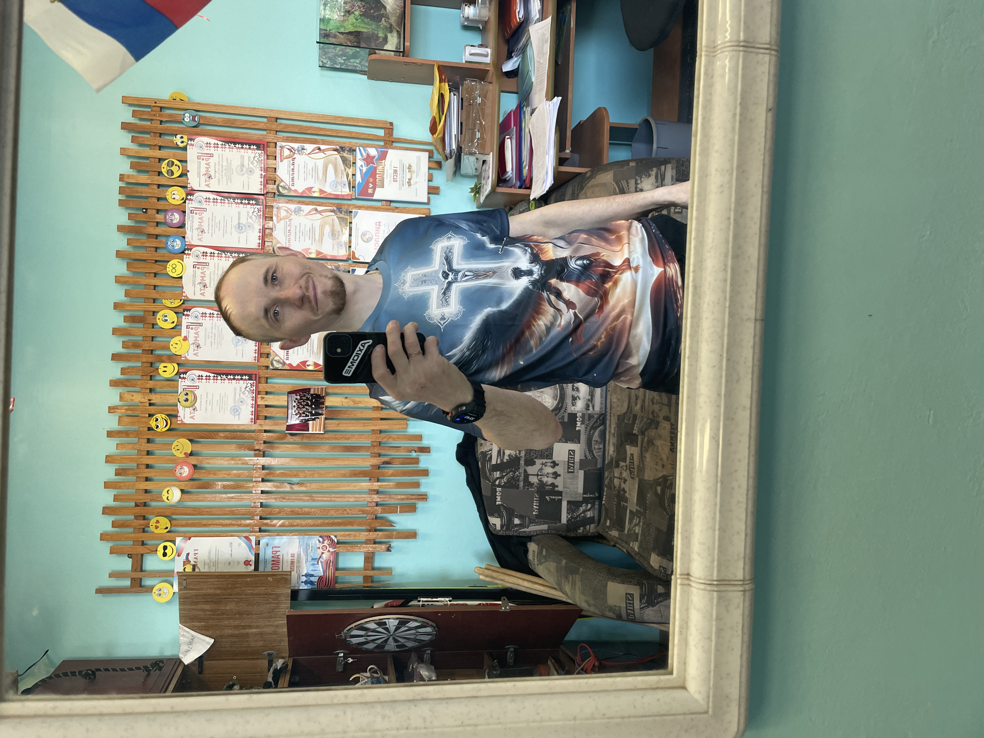 A Caucasian male with thinning hair and a short beard is seen in a mirror reflection, holding a black smartphone with the word "EMOIXA" displayed. He wears a t-shirt with a stylized cross and angel wing graphic. In the background, a wall display features wooden slats adorned with emoji stickers and framed certificates. A Russian flag is visible in the top left corner. Below the display is a wooden cabinet with a dartboard and other miscellaneous items on top. The environment suggests a domestic setting, possibly in the city of Sarapul, Russia, given the presence of the Russian flag. The subject's focus is on taking a selfie in the mirror.