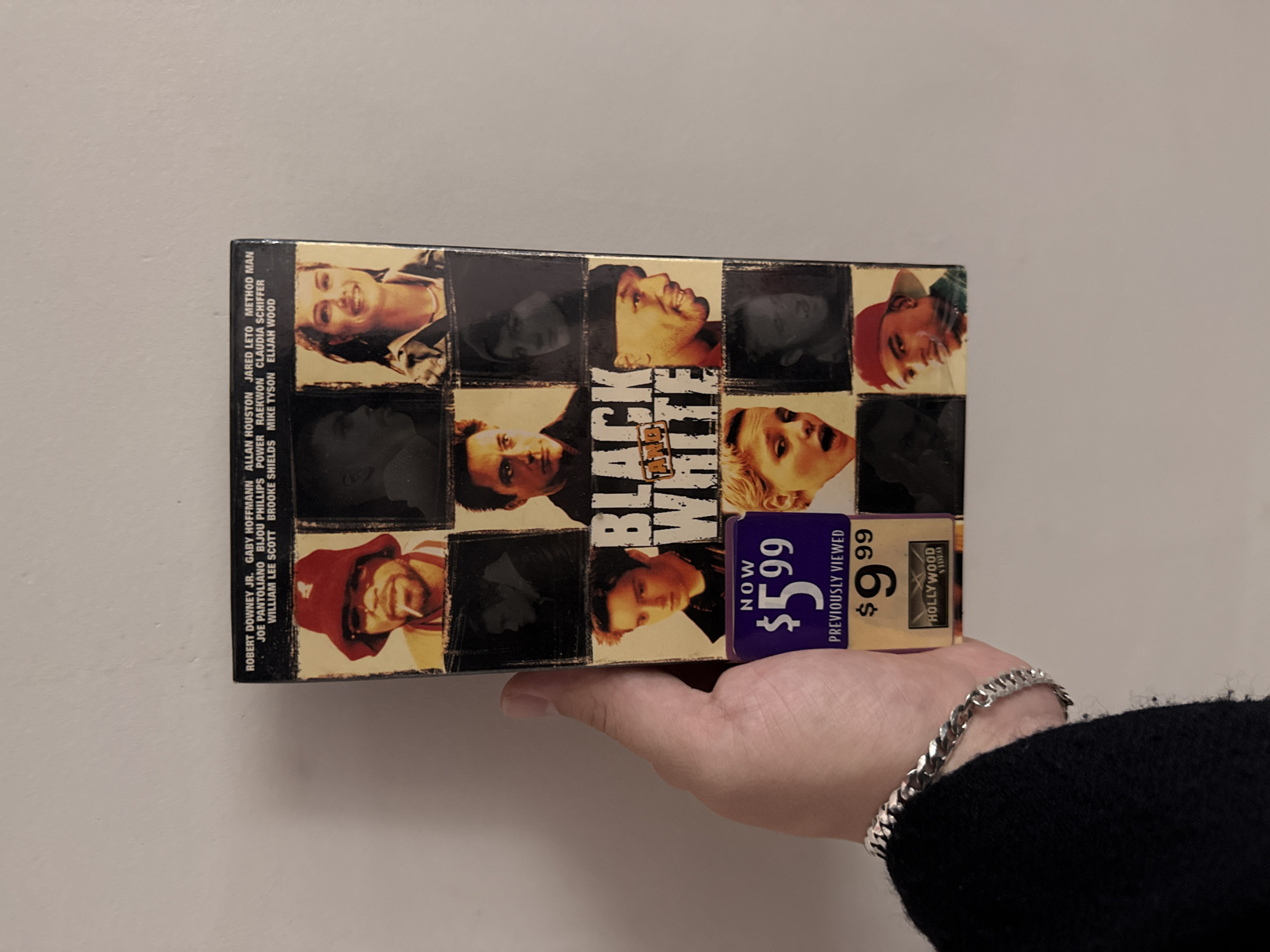 A person holds a VHS tape of the film "Black and White" in their hand. The tape features a checkerboard pattern of headshots on the cover and a Hollywood Video sticker indicating a reduced price of $5.99. The person is wearing a silver bracelet and a black sweater. The background is a plain, light-colored wall.