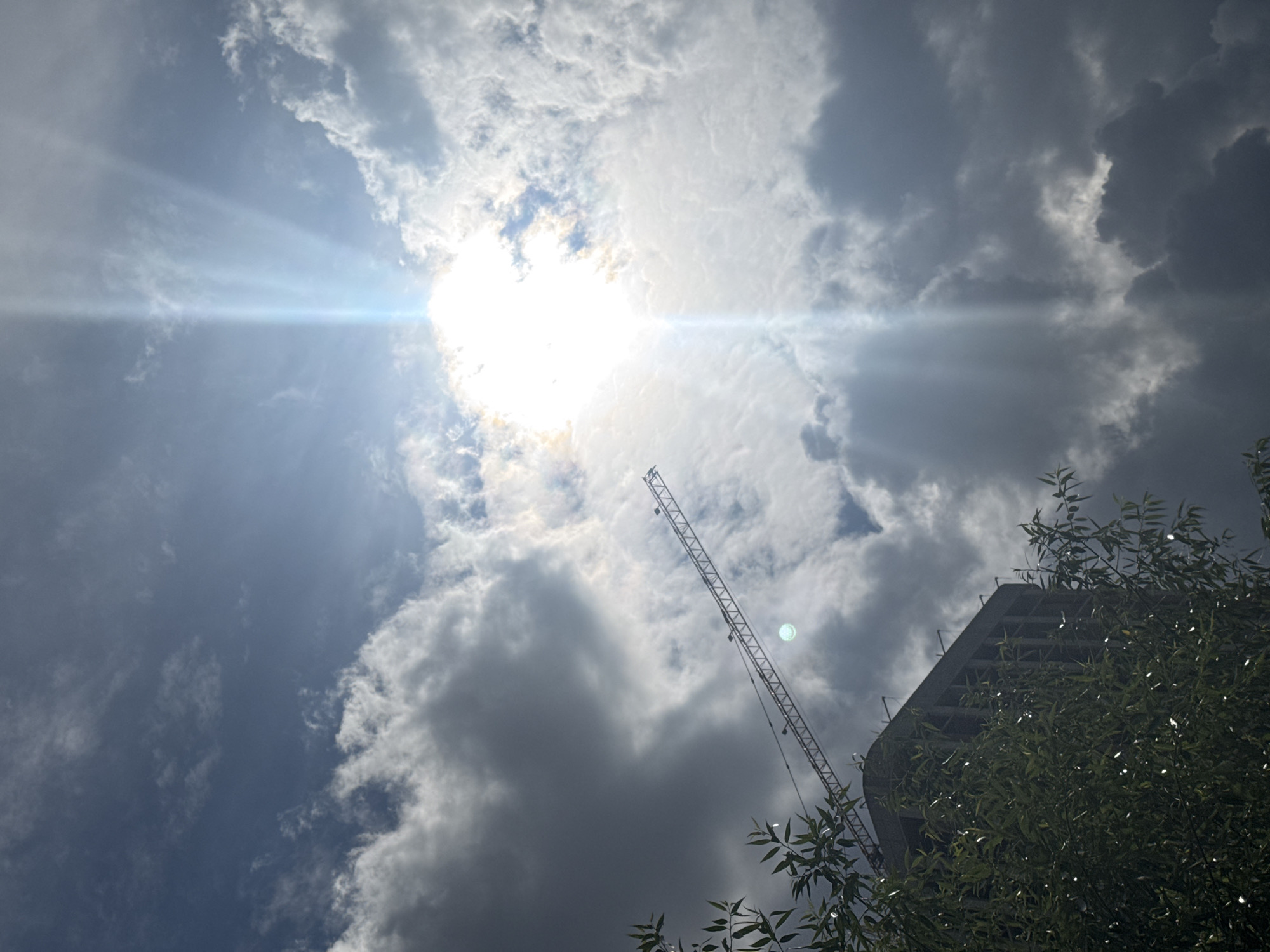 This is a low-angle, wide shot photograph taken from a street-level perspective, looking up towards a partially cloudy sky. The image depicts an urban construction scene. In the foreground, on the left, a portion of a tree with dark green leaves is visible, partially obscuring the view of a tall, unfinished building. The building is made of concrete and appears to be undergoing construction, with visible structural elements. Extending diagonally across the mid-ground from left to right is a construction crane. The sky is a mix of dark, heavy clouds and brighter patches where the sun is shining through. The sun is intensely bright, located in the upper center of the frame, creating a strong lens flare and visible sunbeams. The overall mood is dramatic, with the contrast between the dark clouds and the bright sun, and the presence of construction elements. This scene is located in Izhevsk, Russia.