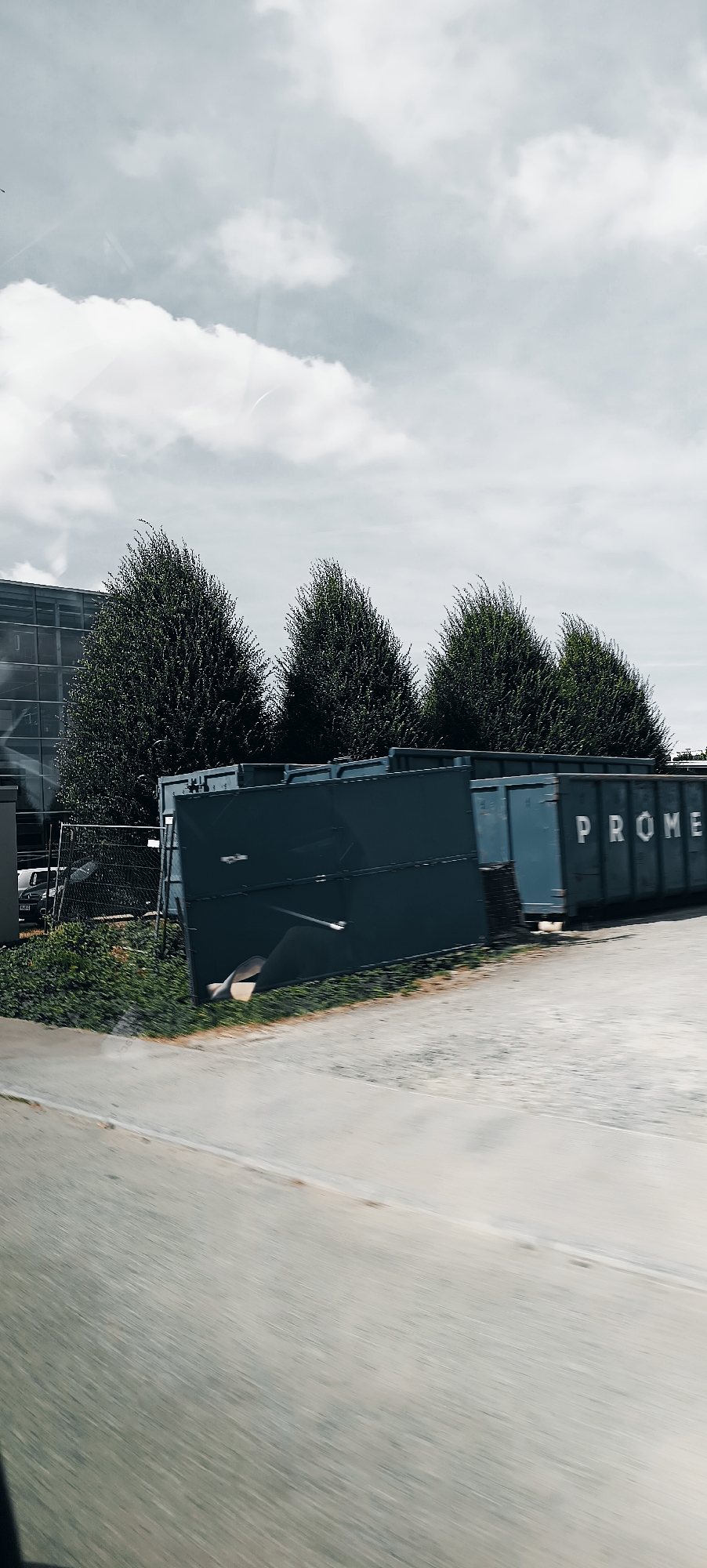 This media file captures an outdoor scene in Fellbach, Germany. The primary objects are several large, dark green industrial dumpsters or roll-off containers positioned in a line. One dumpster clearly displays white lettering spelling out "PROME" on its side. Behind the dumpsters, a row of lush, dark green trees stands tall, some appearing to be manicured. Further in the background, a modern building with a glass facade is visible, reflecting the sky. A chain-link fence runs in front of the trees and the building. The ground in the foreground is a paved area, possibly a parking lot or road, with a grassy strip between it and the dumpsters. The sky is overcast with soft, white clouds, suggesting a daytime scene with diffused light, perhaps mid-morning or afternoon. The overall mood is neutral and industrial. There is no visible activity taking place, implying the image was captured as a static observation. The slight blur in the foreground, particularly on the paved area, suggests the photo may have been taken from a moving vehicle.