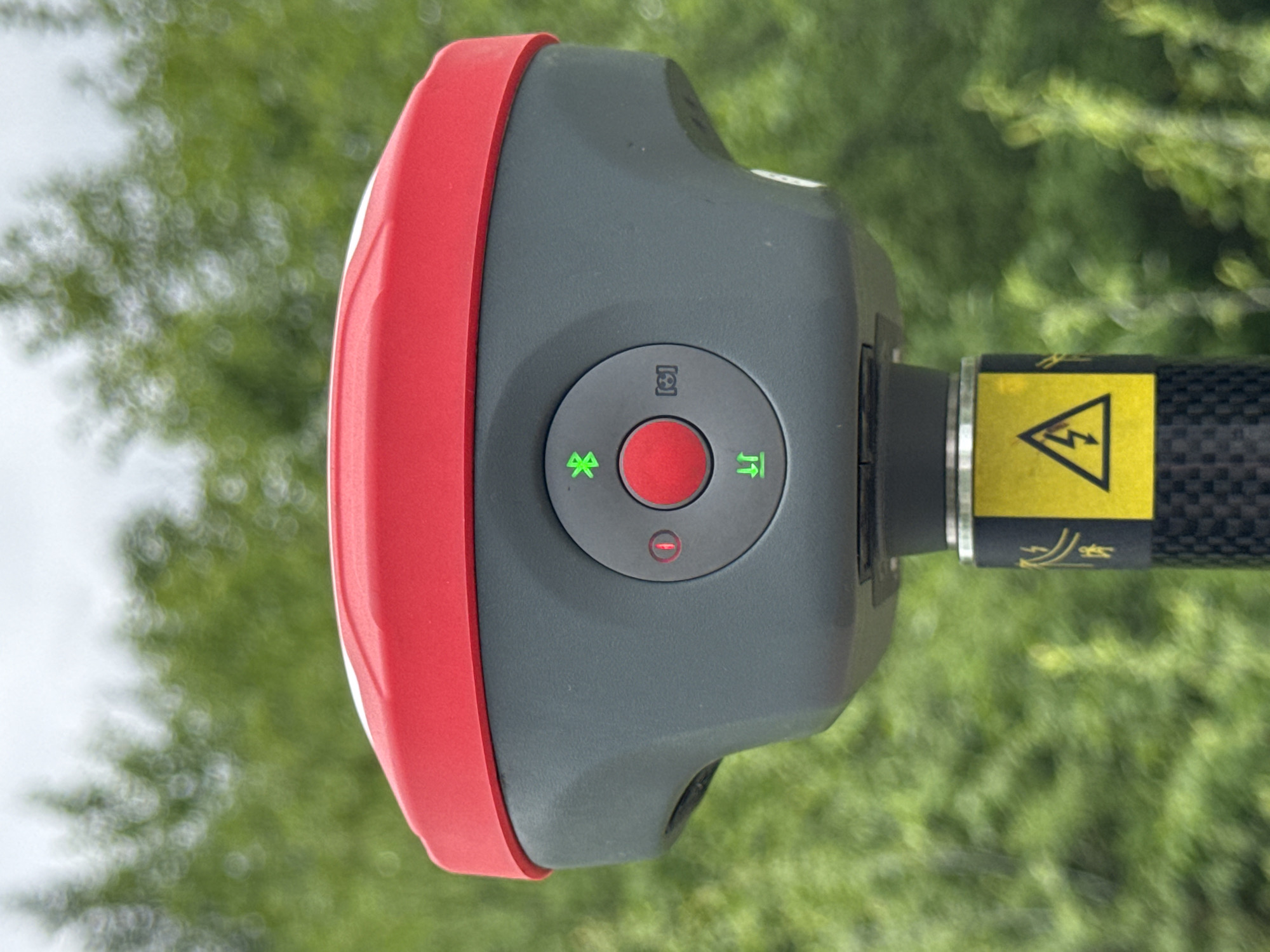 A close-up, eye-level, medium shot features the top portion of a grey and red surveying device mounted on a black carbon fiber pole. The device has a red circular button in the center, surrounded by an array of icons. To the left of the button, a red circle with a horizontal line inside indicates a power button. Above the red button, a green Bluetooth symbol is illuminated. Below the red button, two green arrows point up and down, suggesting a leveling function. To the right of the red button, a grey icon resembling brackets with a central dot is present. A yellow sticker with a black lightning bolt inside a triangle is affixed to the pole directly below the device. The background is heavily blurred, consisting of green foliage under an overcast sky. The scene appears to be outdoors, likely in a natural or semi-rural setting. The city of Murtygit, Russia, is not discernible in the image.