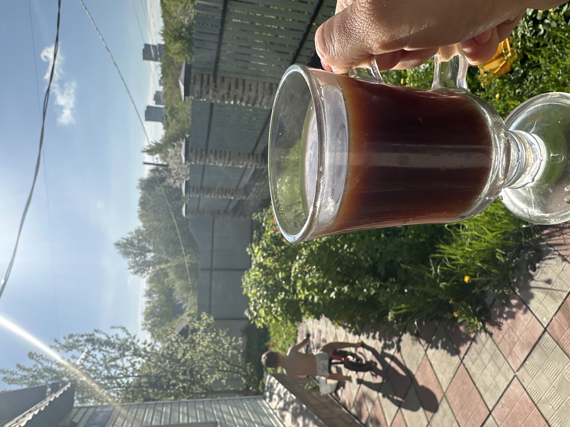 A hand holds a glass mug filled with a dark brown liquid, possibly coffee or tea. The foreground features grass and a tiled patio. In the background, a wooden fence borders the scene, behind which are trees and distant buildings. A shirtless person in shorts is riding a bicycle in the background, partially obscured by shadows and greenery. The photo appears to have been taken outdoors on a sunny day. This scene takes place in Nizhny Novgorod, Russia.