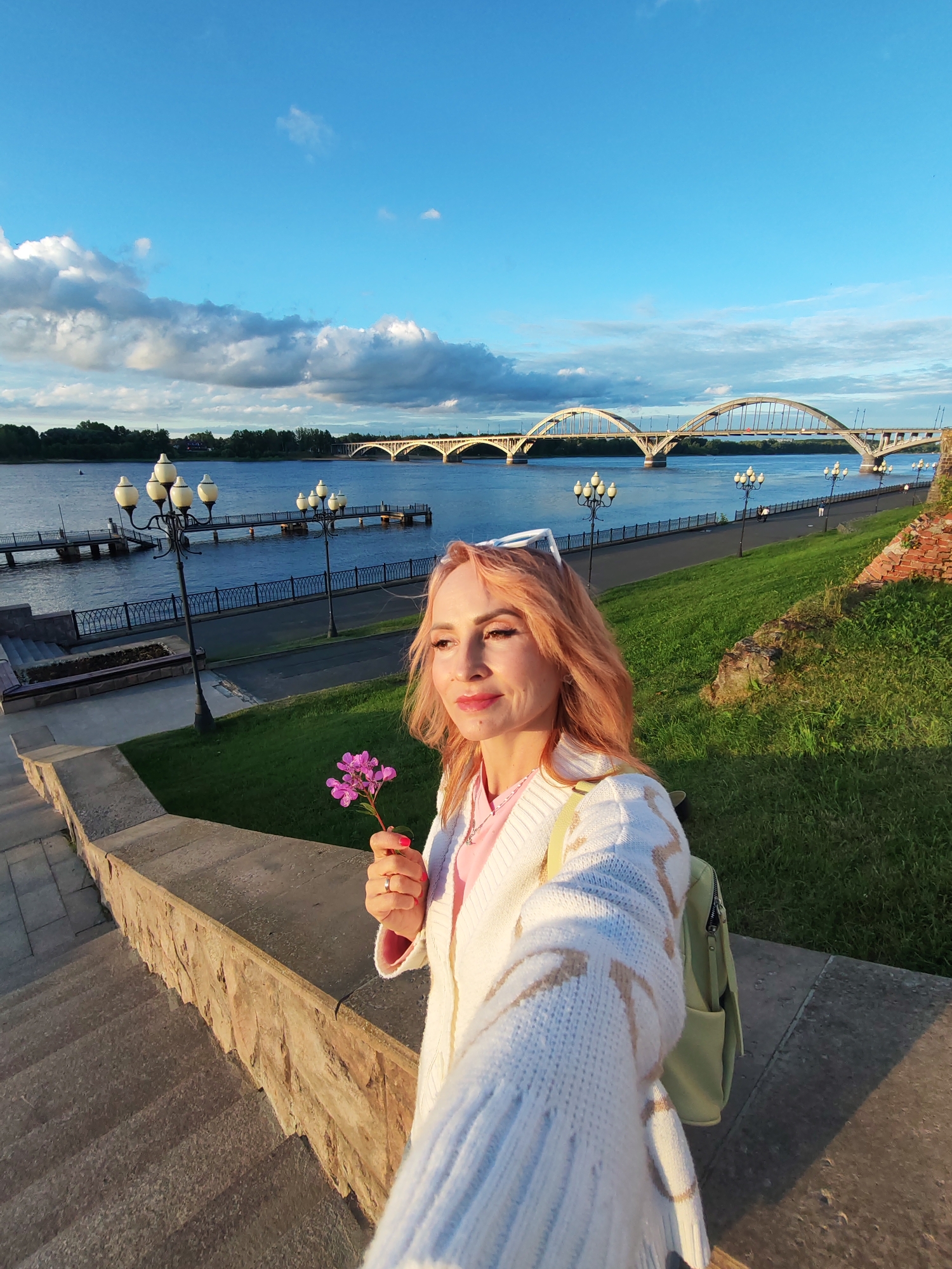 The image depicts a woman holding a purple flower in her left hand, standing on a stone staircase overlooking a wide river. She has fair skin, pink-tinted hair, and is wearing a white knitted cardigan over a pink shirt. A light green backpack is visible on her right side. In the background, a long, multi-arched bridge spans the river, with a city skyline and trees lining the opposite bank. A waterfront promenade with lampposts runs along the near side of the river. The sky is a vibrant blue with scattered clouds, indicating a clear day. The scene appears to be a public outdoor space, likely a park or embankment, in the city of Bol'shie Mkhi, Russia. The lighting suggests it is late afternoon or early evening.