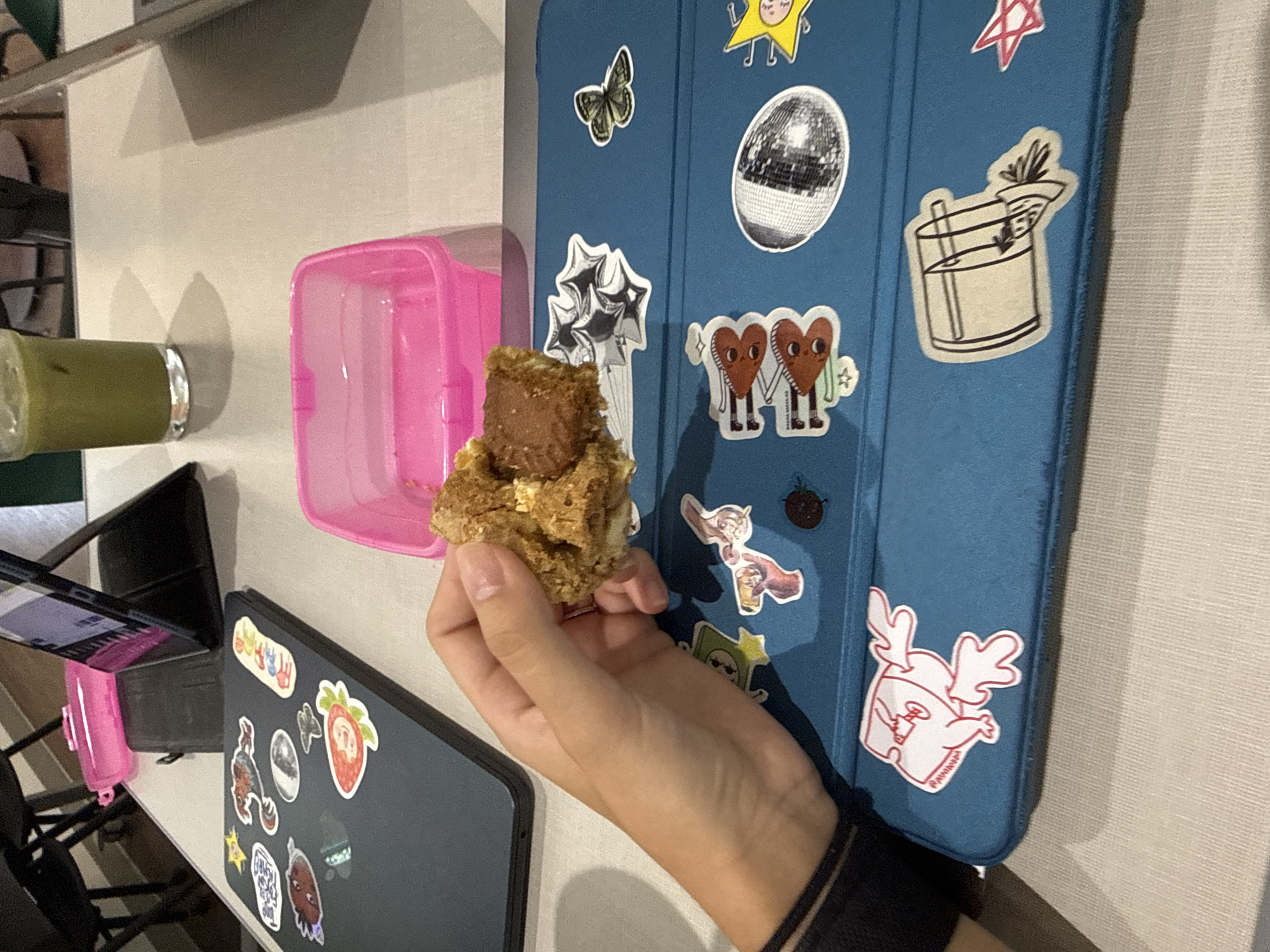 The image, captured in San Andrés Cholula, Mexico, presents a close-up, tilted view of a person's hand holding a crumbly, brown food item, possibly a cookie or a granola bar, with a darker brown layer on top. The hand has light skin and neat, natural nails.
The scene is an indoor, casual setting, likely a desk or table. Dominating the right side of the frame is a blue tablet or laptop case, extensively decorated with various stickers. These stickers depict a silver butterfly, a disco ball, a yellow star with a face, a stylized cocktail glass with a straw, two cartoon heart-shaped figures holding hands with the partial text "PREMA MATES," a cluster of silver star-shaped balloons, and a white cartoon character with pink antlers and the text "@PIGGYDUCK."
To the left of the hand, a bright pink, empty plastic container is visible. Further left, a clear glass with a metallic rim contains a green liquid, presumably a smoothie or juice. In the lower left foreground, a corner of a black laptop or tablet is seen, also adorned with stickers, including a red strawberry and colorful handprints. A small pink stand is positioned next to this device. The background is softly blurred, showing glimpses of furniture and a person's leg or clothing. The overall impression is a relaxed personal space, possibly during a snack break. The time of day is not evident, given the indoor lighting.