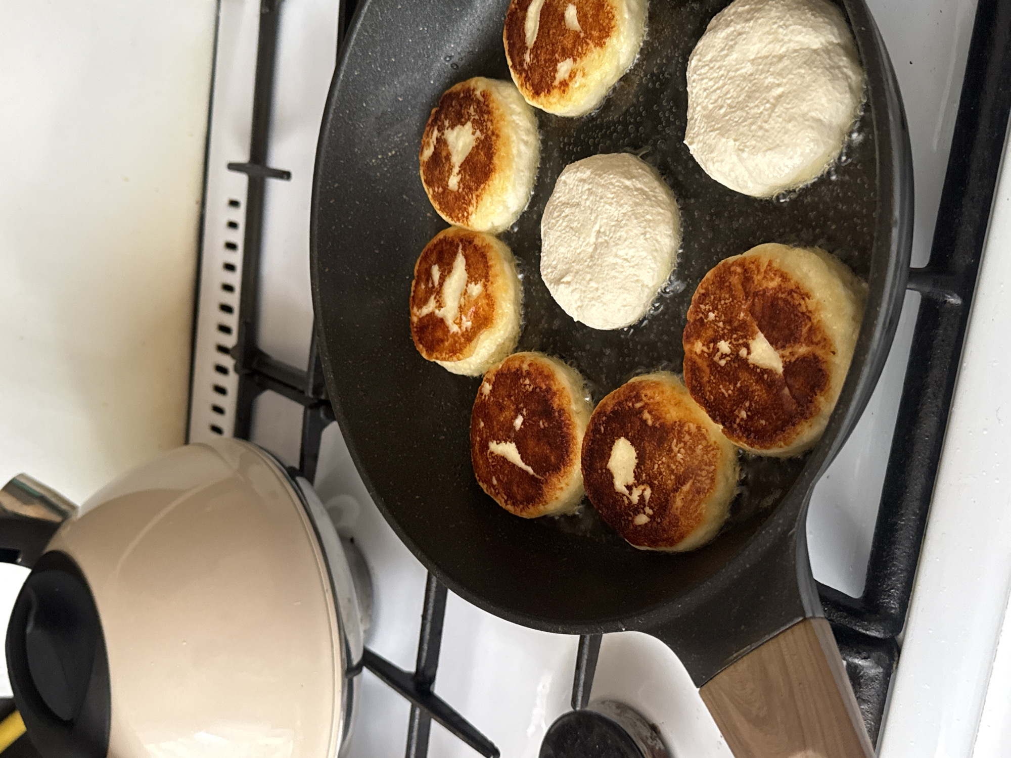 A top-down, close-up shot captures syrniki, small, round cheese pancakes, being fried in a black, non-stick pan on a white gas stovetop. The pan is filled with sizzling oil, and several syrniki are in various stages of cooking. Some are golden brown and slightly puffed, indicating they are nearly done, while others are pale and appear to have just been placed in the pan. The syrniki have a fluffy, slightly irregular texture.
To the left of the pan, a portion of a light-colored electric kettle with a black handle is visible. The background consists of the white stovetop and black grates. The scene suggests a domestic setting, likely a kitchen, in Chaikovsky, Russia, during the daytime. There are no people visible, and the focus is entirely on the food preparation. The overall impression is one of a home-cooked meal in progress.