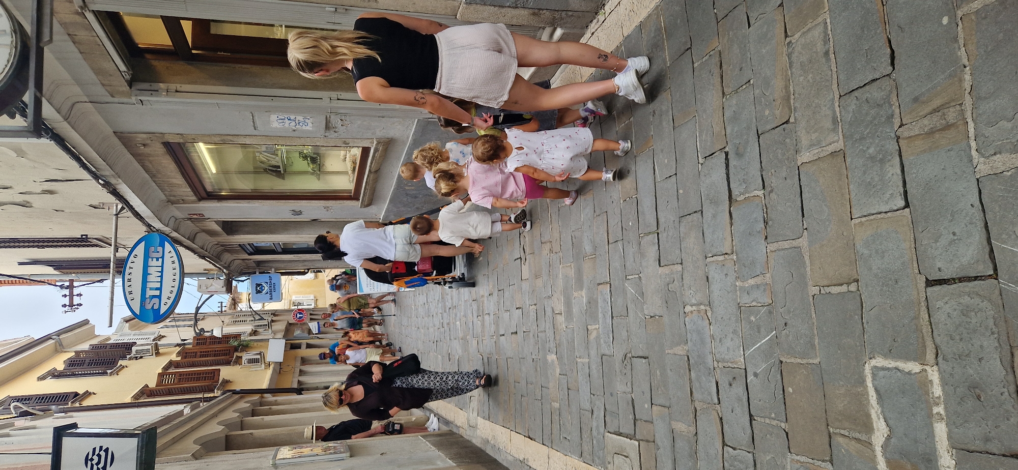 In Koper/Capodistria, Slovenia, a group of people walk down a narrow, cobblestone street lined with old buildings. In the foreground, a blonde woman in a black tank top and light-colored shorts walks away from the camera, her back to the viewer, with a tattoo on her left arm. She is followed by a line of young children, some walking and some being pushed in a stroller. The children are dressed in various colorful outfits. Behind them, a woman in a white t-shirt and shorts pushes the stroller. Further down the street, other people are walking, including a woman in a dark jacket and patterned pants carrying a large bag. Signs for "URARSTVO STIMEC OROLOGERIA" and "BARBERSHOP KOPER" are visible on the buildings. The architecture suggests an older European city.
