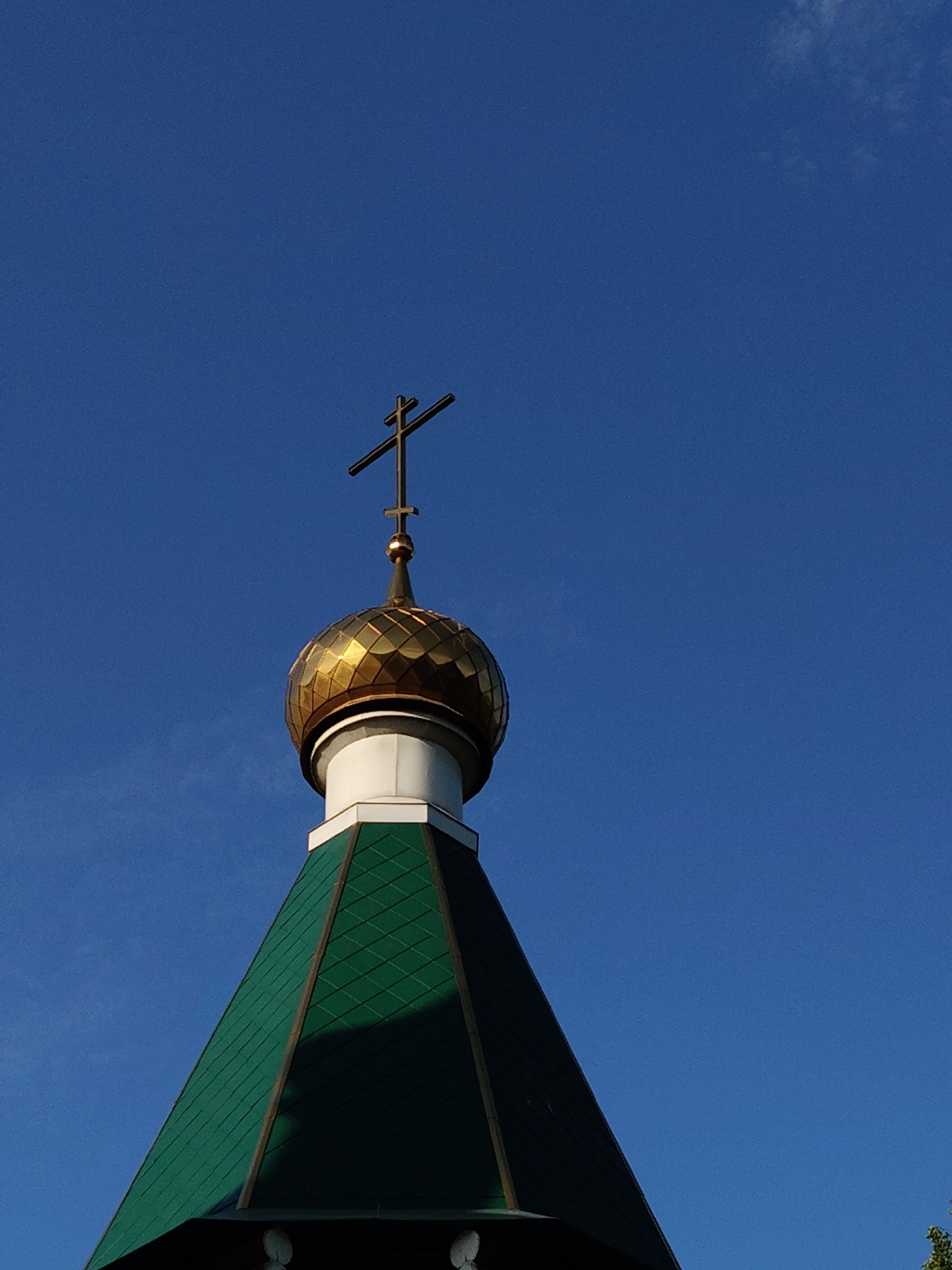 The image shows the top of a church with a green roof, a white middle section, and a golden dome topped with a black cross. The background is a clear blue sky with a few barely visible clouds. The photo appears to be taken in daylight. The city and exact location are unknown.