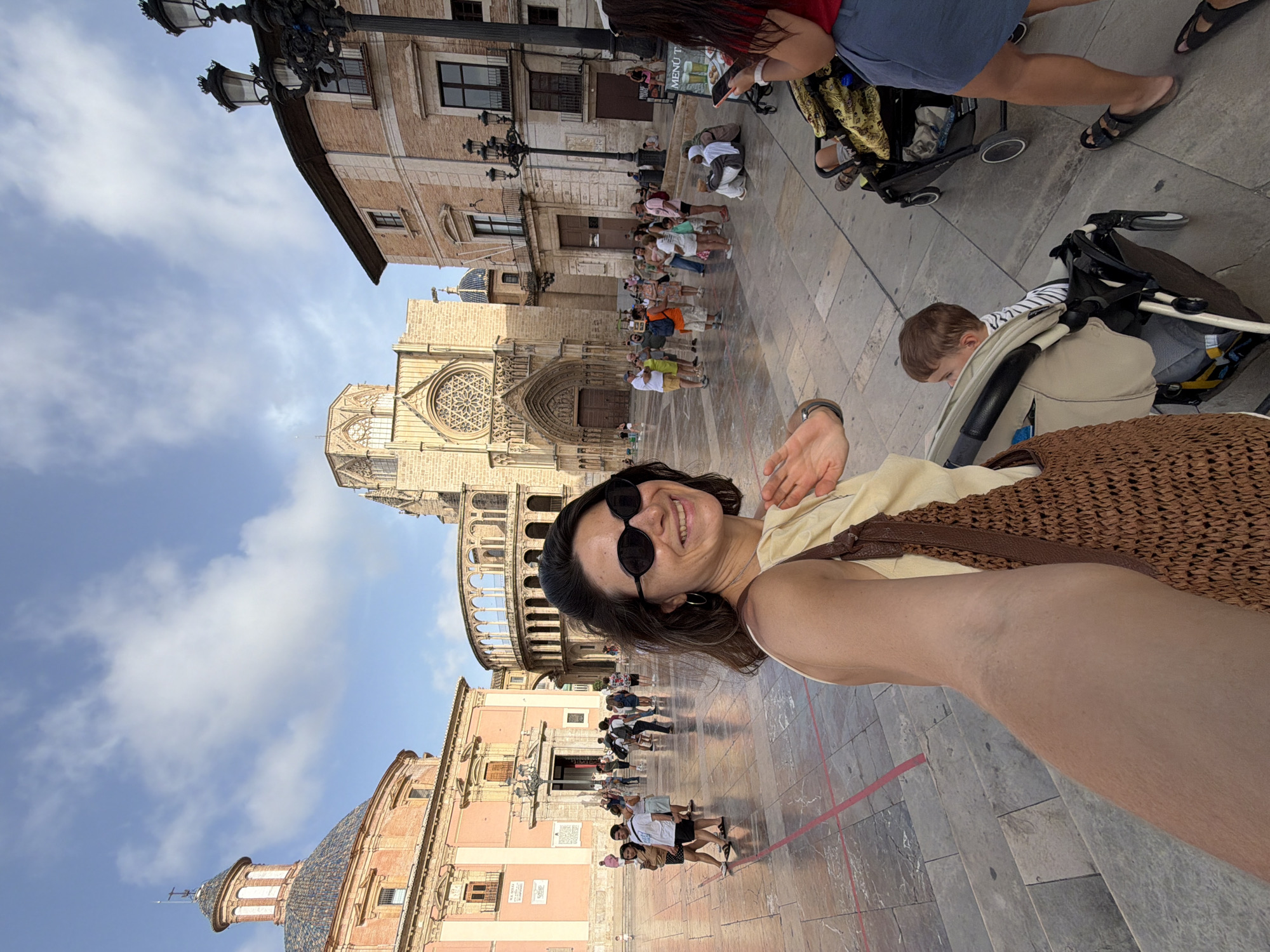 In an outdoor plaza, a woman with dark hair, wearing large dark sunglasses and a beige off-the-shoulder top, takes a selfie. She holds a woven brown bag over her shoulder and waves with her left hand. Behind her, several people are walking and gathered in small groups. In the background, a large Gothic cathedral with a rose window and ornate architecture dominates the scene, along with other historic buildings and a street lamp. A baby in a stroller is visible in the foreground to the right of the woman. The ground is paved with large square tiles. The sky is blue with scattered white clouds.
