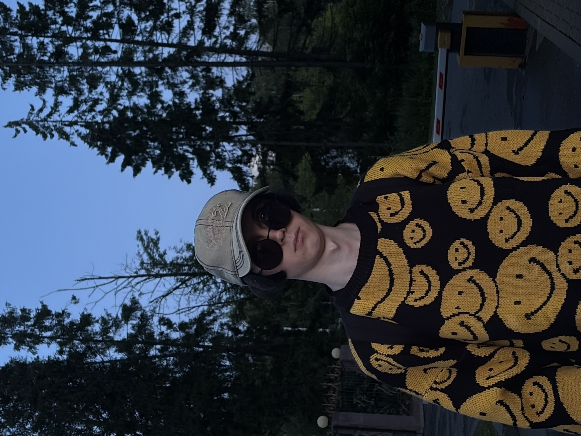A young person stands outside in front of trees. They are wearing a tan baseball cap, round black sunglasses, and a black sweater with yellow smiley faces. A black backpack is visible over their shoulders. In the background, there is a metal fence with decorative spheres, a road, and a yellow and gray barrier gate. The sky is a dim blue, suggesting it is either dawn or dusk.