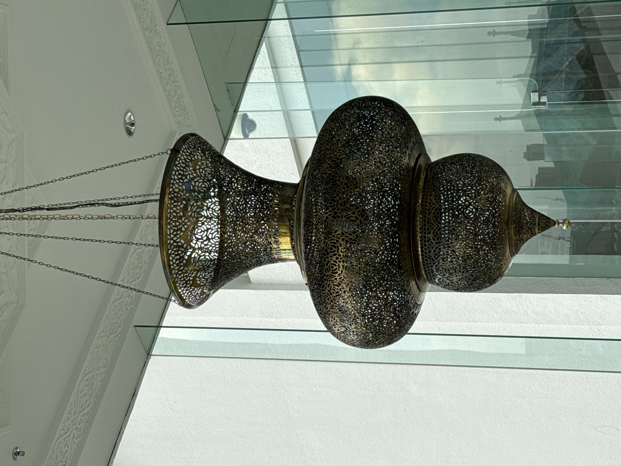 A detailed, low-angle shot captures an ornate, pierced metal chandelier hanging in a modern architectural setting, possibly an interior space in Kuala Lumpur, Malaysia, given the architectural style.
The chandelier is the focal point, exhibiting a complex, pierced metalwork design. It consists of multiple spherical and flared sections joined by smooth metallic bands. The perforated metal allows light to pass through, creating intricate patterns. The chandelier is suspended by multiple chains.
The background features a modern, clean aesthetic with white walls, geometric architectural details, and glass panels. The glass reflects the city landscape outside, offering a glimpse of buildings and sky. The light reflects off the glass surfaces, creating a bright and airy atmosphere. A recessed light fixture is also visible in the ceiling.