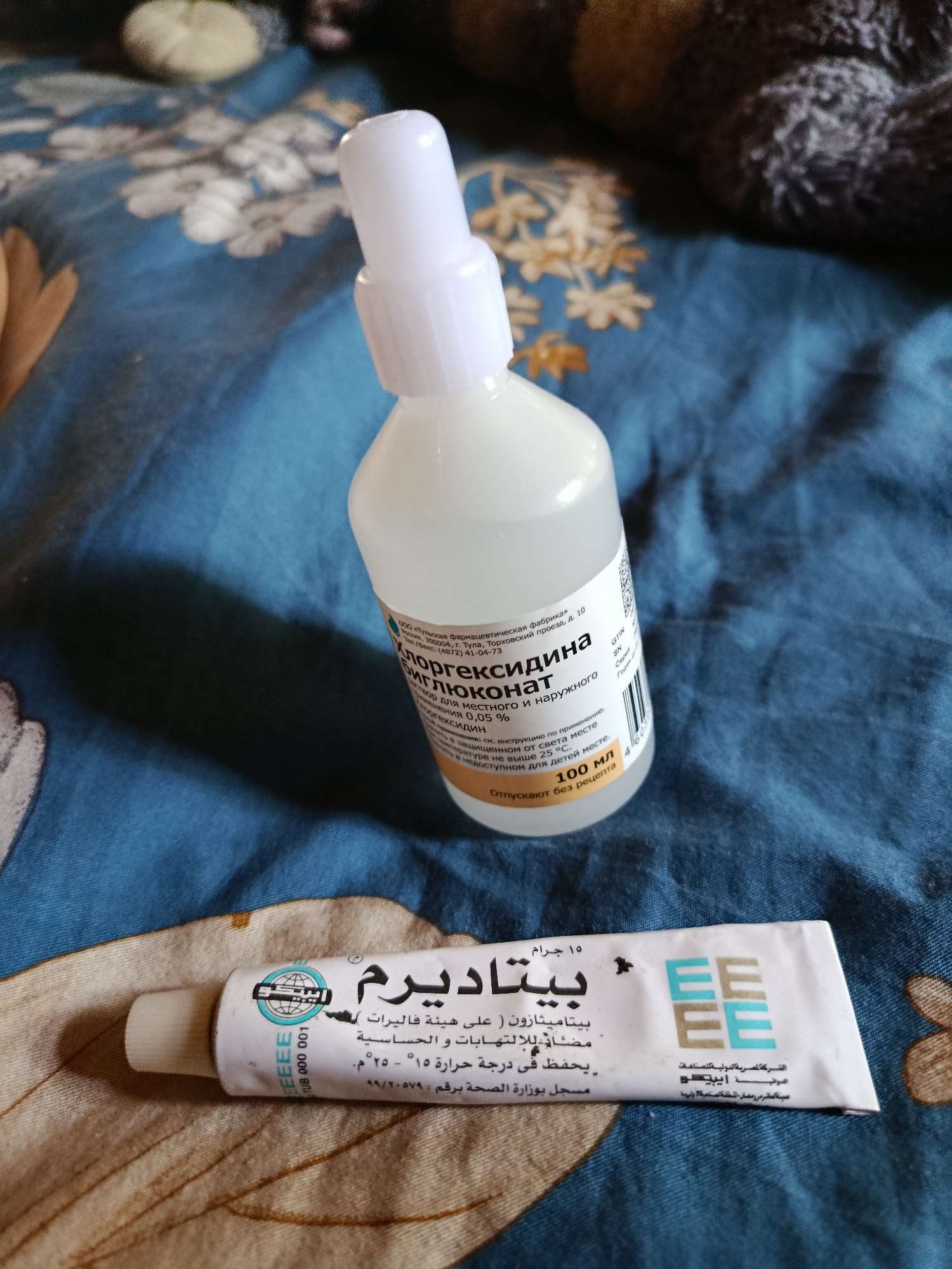 The image displays two pharmaceutical products placed on a blue fabric surface adorned with a white and light brown floral pattern. In the upper center, a white plastic bottle with a spray top is visible. The bottle's label, written in Cyrillic, identifies its contents as "ХЛОРГЕКСИДИНА БИГЛЮКОНАТ" (Chlorhexidine Bigluconate) solution, 0.05% for local and external use, in a 100 ML volume. The manufacturer is listed as OOO "Tula Pharmaceutical Factory," located at Torkhovsky proezd, bld. 10, Tula, Russia. The label also indicates it is available without a prescription and should be stored below 25°C, protected from light and out of reach of children.
At the bottom of the frame, a white tube of cream or ointment lies horizontally. Its label, primarily in Arabic with some English, identifies the product as "بيتاديرم" (Betaderm), containing 15 grams of Betamethasone. It is described as an anti-inflammatory and anti-allergic, to be stored at 15-25°C. The manufacturer is "الشركة المصرية الدولية للصناعات الدوائية" (The Egyptian International Pharmaceutical Industrial Company), also known as EIPICO, with a logo showing four 'E's. The tube's registration number with the Ministry of Health is 59/2079. The background is out of focus, indicating an indoor setting. No individuals are present in the scene.