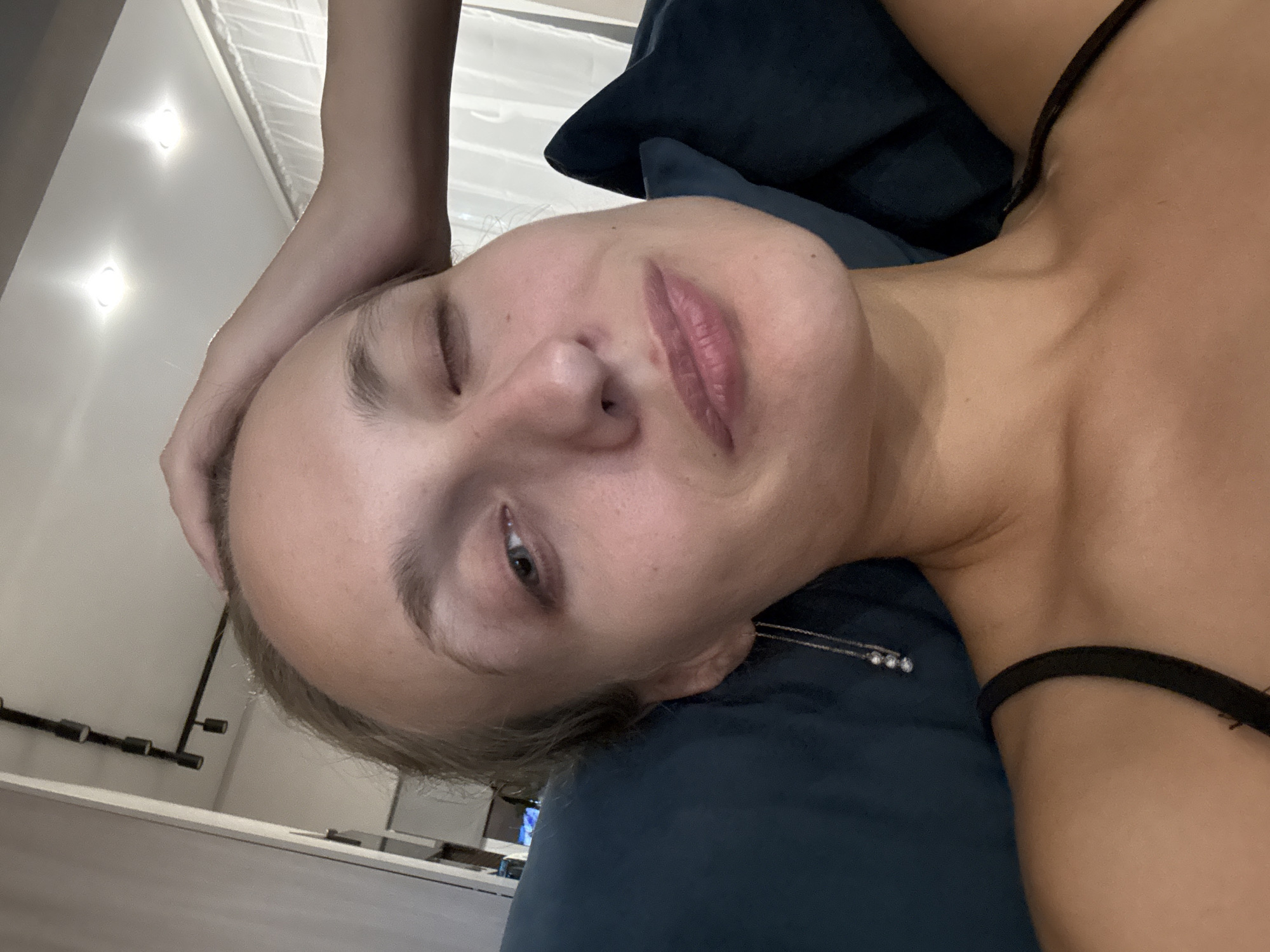 In an indoor, dimly lit environment, a young Caucasian woman is captured in a close-up, selfie-style shot. She is lying down, with her head resting on a dark blue pillow and her right arm extended behind her head, appearing to be touching her hair. Her left eye is open, while her right eye is closed in a wink. She is wearing a black tank top, and a long, delicate earring with three small, pearlescent beads hangs from her left ear. The background suggests a domestic setting, with part of a wall and a ceiling with track lighting visible. There are no specific indicators to place the scene in the city of Подшивалово, Russia.