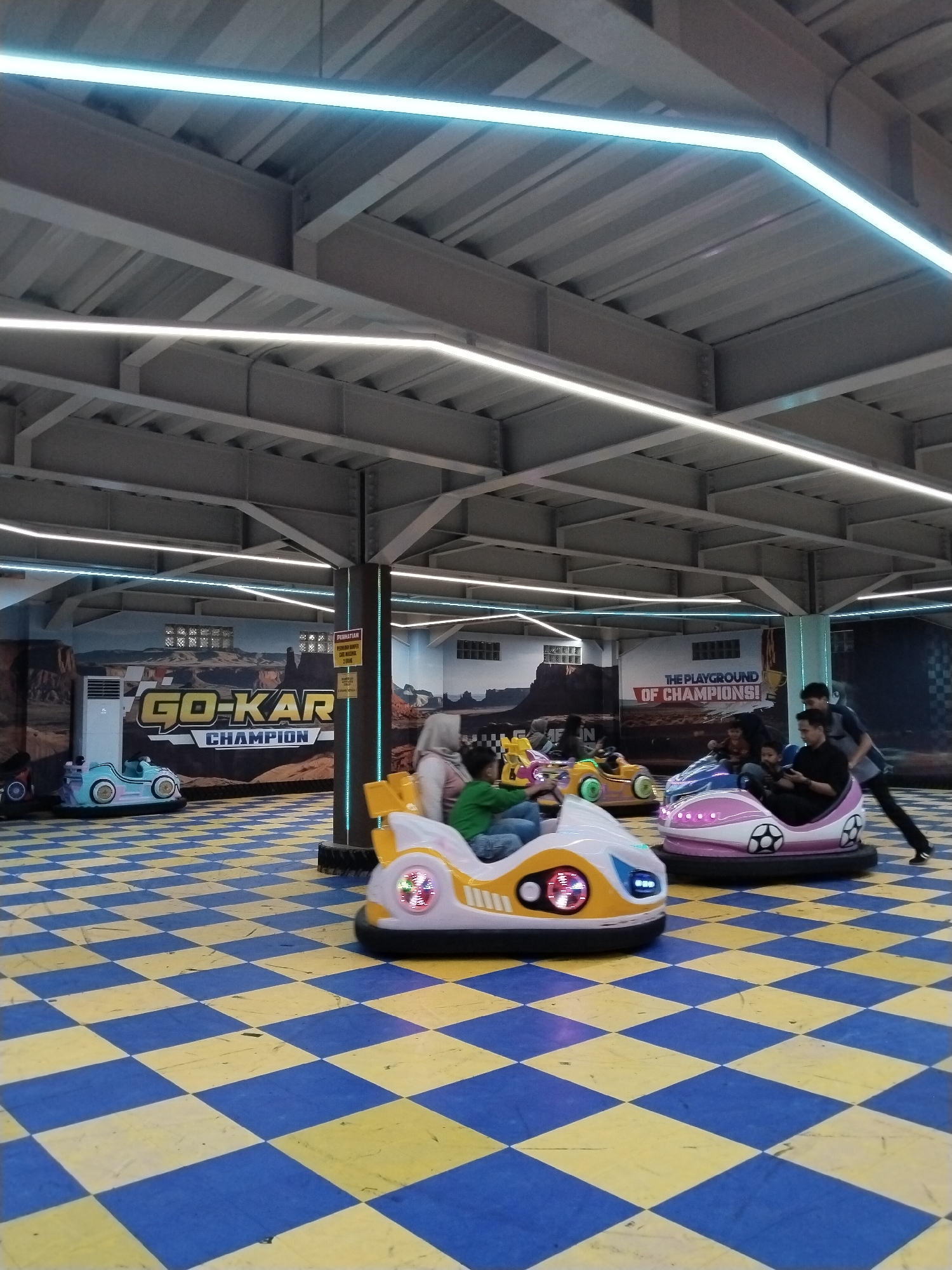 This image depicts an indoor amusement park, likely a bumper car arena, located in Kecamatan Pati, Indonesia. The floor is a checkerboard pattern of blue and yellow tiles. Several bumper cars are visible, some occupied by individuals. In the foreground, a white and yellow bumper car with LED lights is in motion. To the right, a pink and white bumper car with a soccer ball motif is also present, with two people inside. In the background, a mural advertises "GO-KART CHAMPION" and "THE PLAYGROUND OF CHAMPIONS". The ceiling is supported by metal beams and illuminated by strip lights. One person, a male wearing a dark shirt and black pants, is walking away from the bumper cars towards the right edge of the frame. Another male, wearing a blue shirt and black pants, is visible behind him.