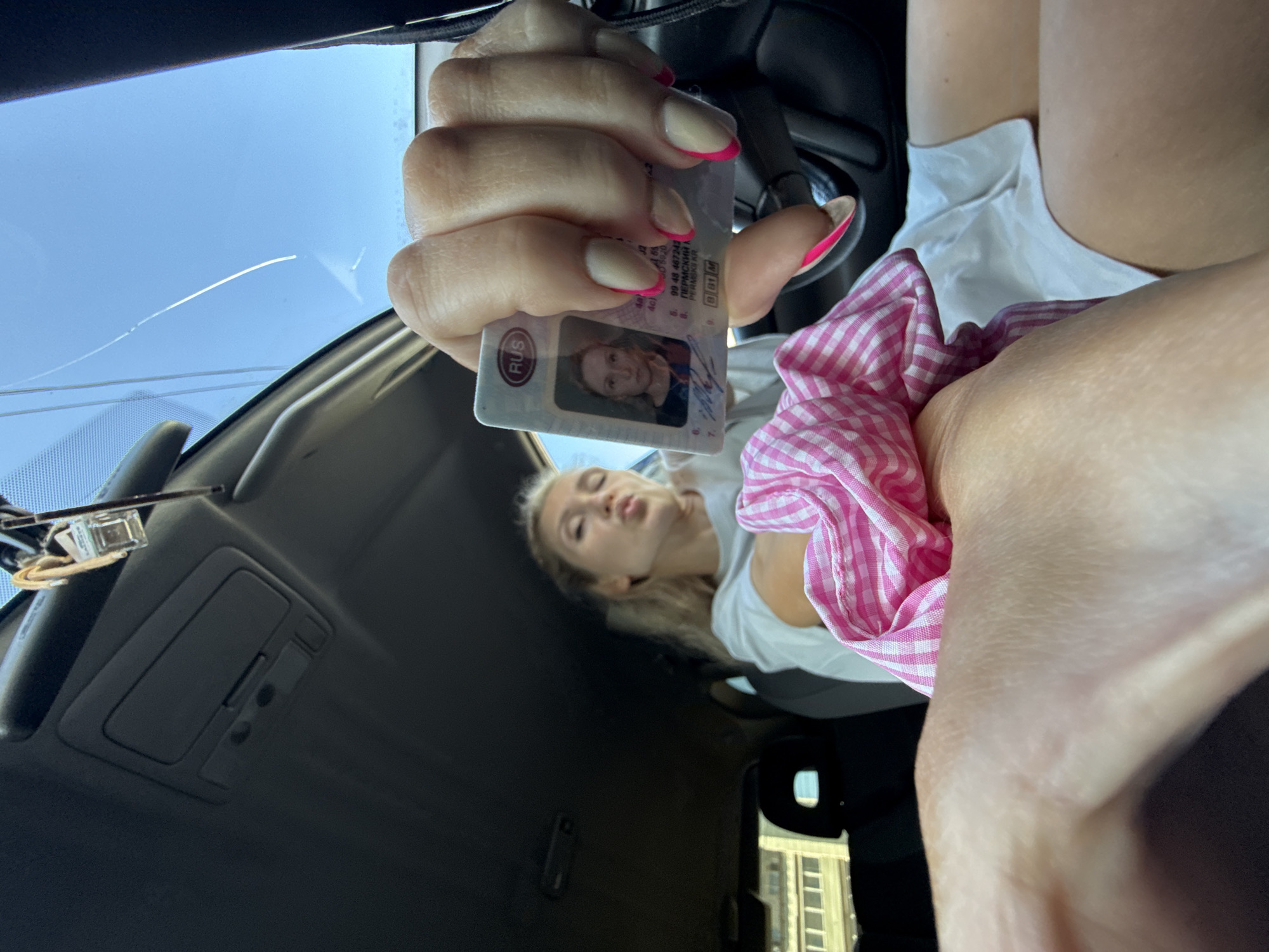 The image shows a person sitting in a car, holding up a Russian driver's license in their hand. The license features a photo of a woman, and the card is marked with "RUS" and mentions "PERMSKI KR," which suggests the Perm region of Russia. A pink and white checkered scrunchie is visible near the person's arm. Through the car windshield, the sky is visible. It is difficult to pinpoint the exact location in Chaikovsky based on the image alone.