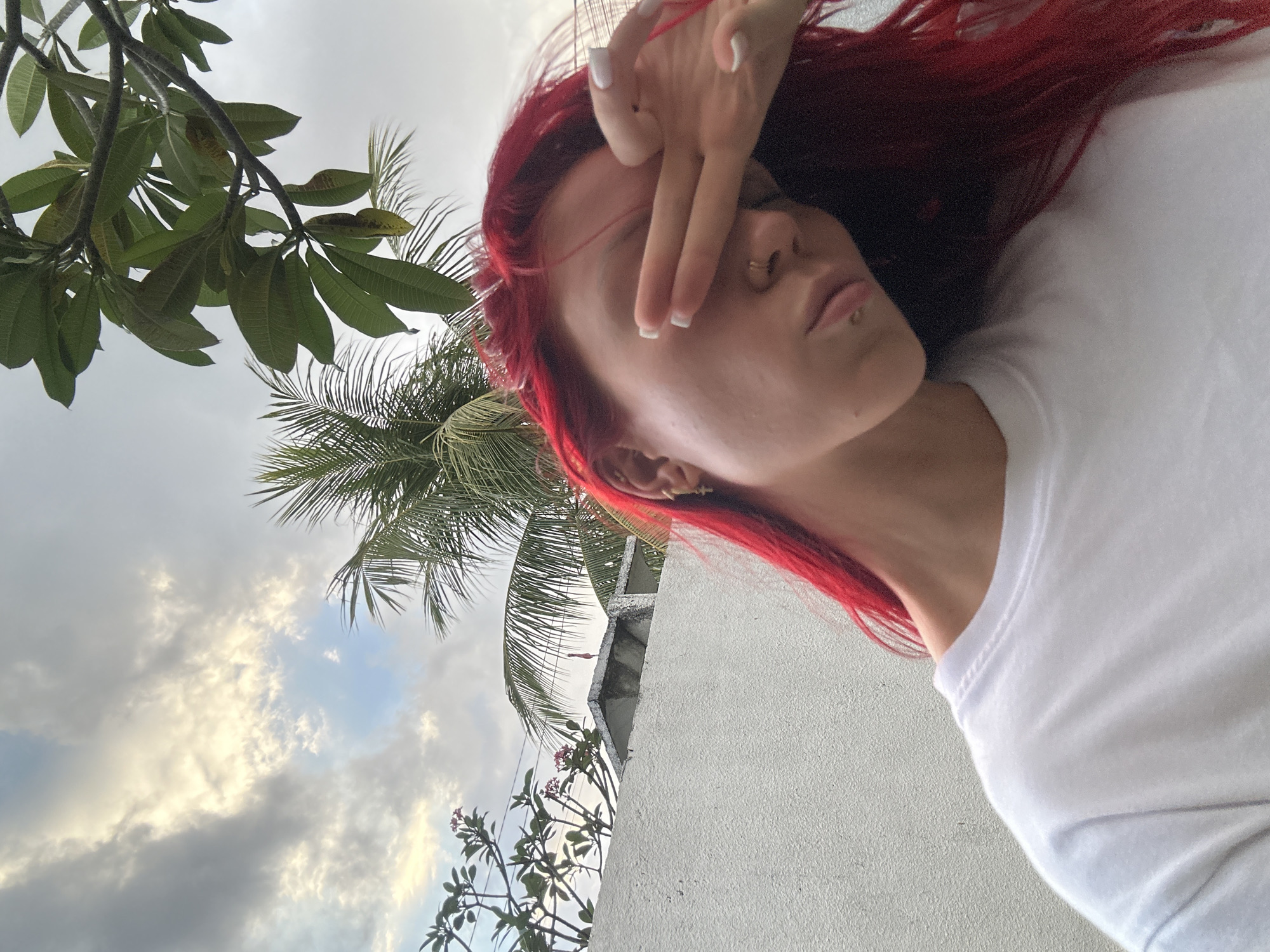 A close-up, slightly Dutch-angled photograph captures a person with vibrant red hair, fair skin, and a white t-shirt, posing outdoors, likely for a selfie. The individual wears a subtle gold-colored nose ring and a small piercing on their lower lip. Their right hand is raised, partially covering their eyes and forehead, with two fingers (index and middle) extended, resembling a peace sign. The fingers display white manicured nails with a French tip.
The background features a blend of natural tropical elements and a simple white architectural structure. Lush green foliage, including broad-leafed branches, possibly Plumeria or Frangipani, is visible in the upper left, while the fronds of a palm tree span across the middle and right sections of the image. A textured white wall or building occupies the lower right quadrant. The sky, seen between the leaves and above the wall, is partially cloudy with patches of lighter, brighter sky, indicating a daytime setting. The overall lighting suggests a bright but somewhat overcast day. The scene is consistent with a tropical location such as North Kuta, Indonesia. There is no visible text.