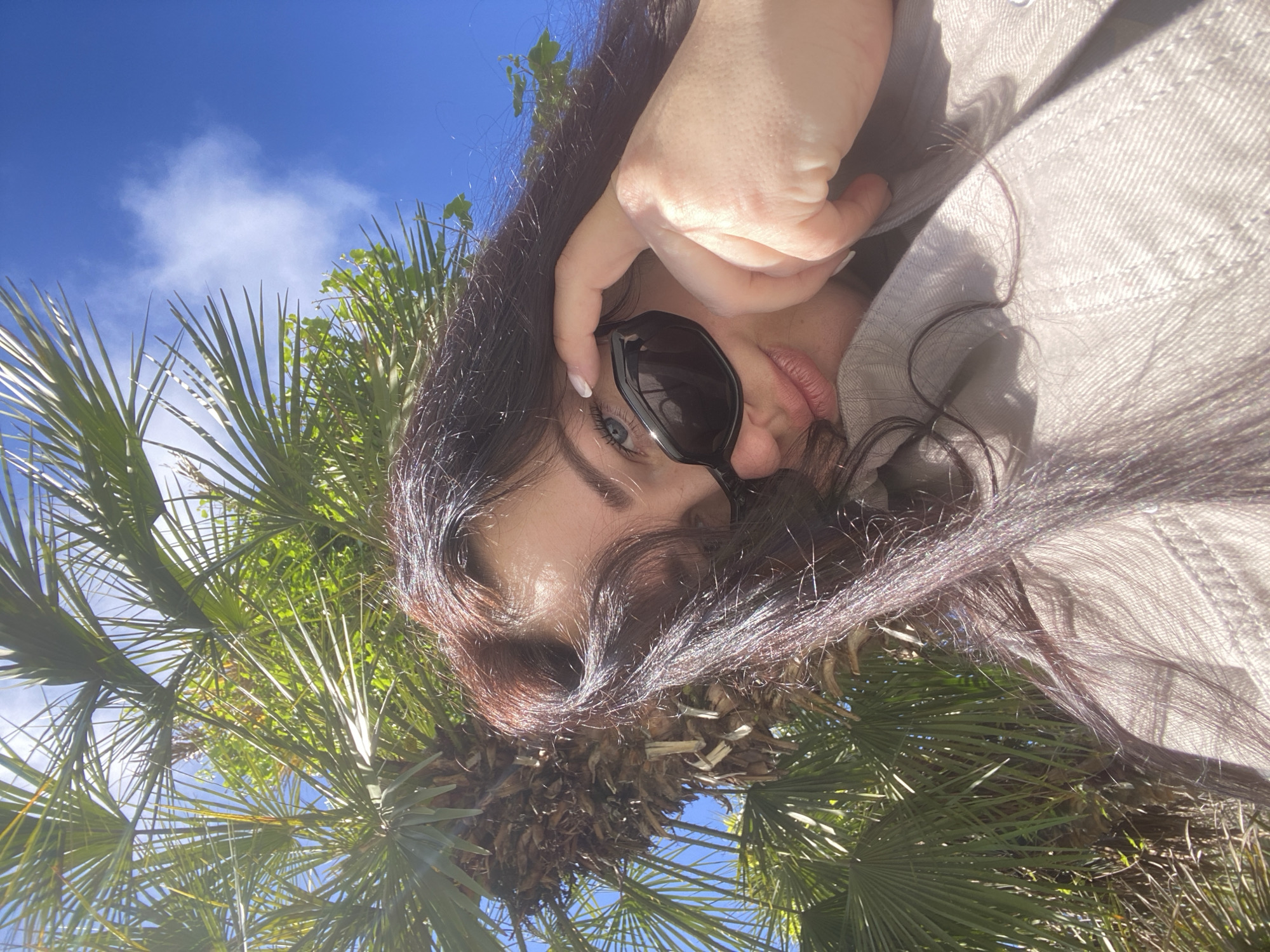 A selfie taken outdoors from a low angle, capturing a person's face, hand, and portions of their clothing with a background of palm trees and a bright blue sky with some white clouds. The person is holding the frame of their sunglasses near their eye. Their hair is dark brown with auburn highlights. The person is wearing a light-colored jacket or shirt. The image is taken from a perspective that looks upward into the canopy of a palm tree. The environment suggests a sunny day in a warm climate, potentially near a coastal area. The specific location in Sochi, Russia is not evident from the image.