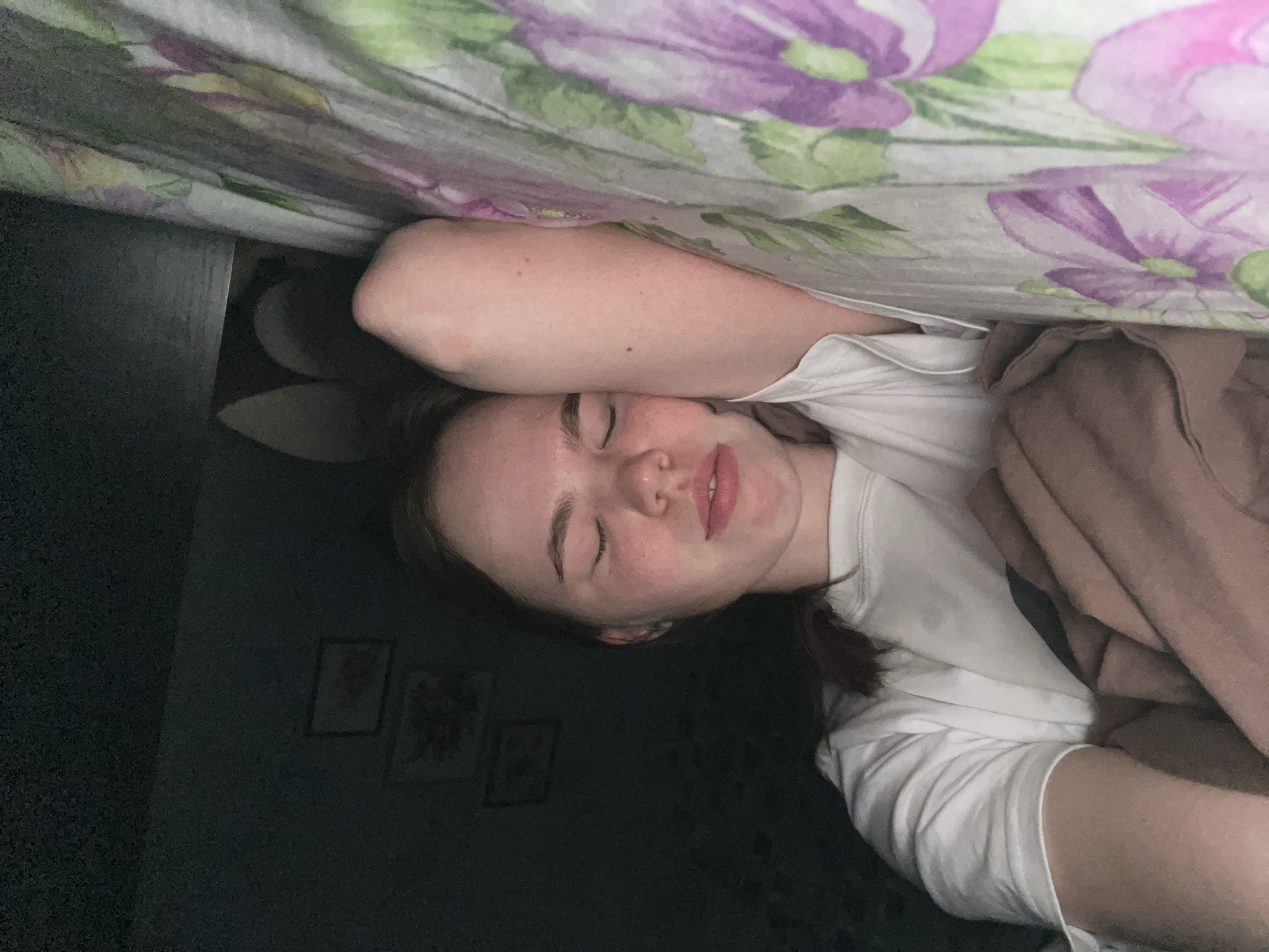 The image depicts a young female asleep or resting in a bed within a room in the city of Chaikovsky, Russia. She is positioned on her right side, with her eyes closed and her head resting on her right forearm. Her lips are slightly parted. She has fair skin, a visible freckle or mole on her right arm, and wears a white short-sleeved t-shirt. A light brown or beige blanket partially covers her upper torso. The bed is fitted with a sheet featuring a prominent floral pattern of large purple and pink flowers with green leaves. The background is dimly lit and dark, showing a wall on which two framed, dark-toned pictures or artworks are faintly visible.