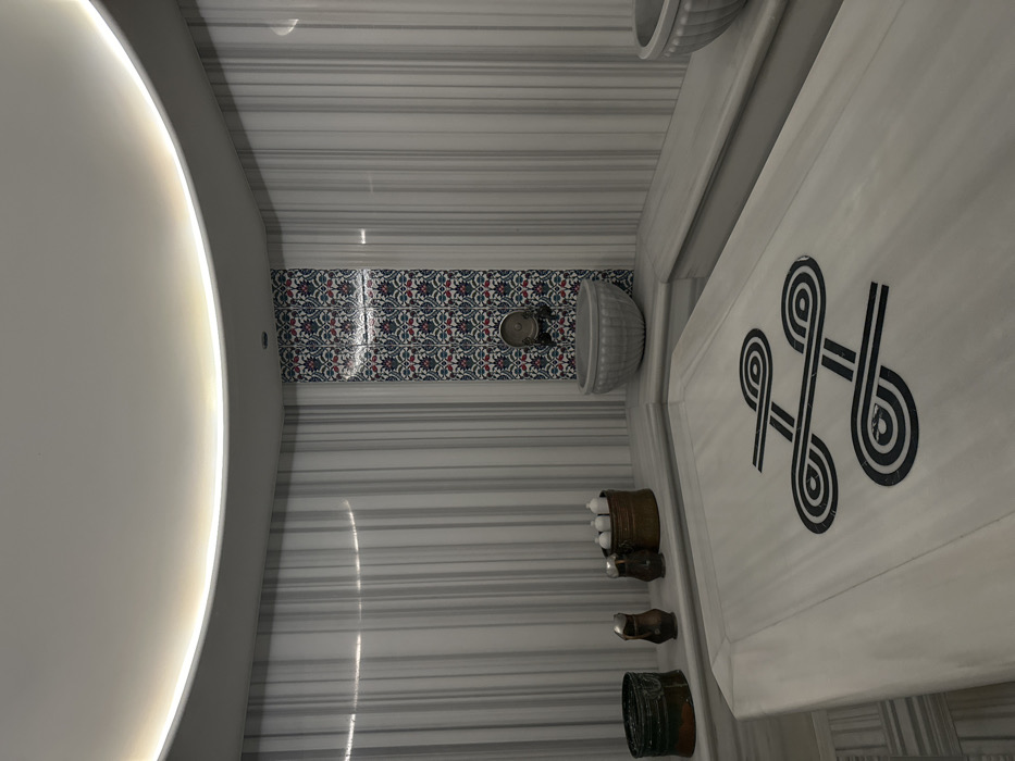 The image depicts the interior of a modern hammam or spa facility located in Bartula, Montenegro. The room features walls and built-in elements constructed primarily from light-colored marble with vertical grey veining. The ceiling is white, curved, and illuminated by concealed LED strip lighting in a cove.
In the foreground, a large, rectangular white marble slab, likely a warming or massage table, occupies the central space. This slab features an intricate black and white geometric knot design inlaid into its surface.
Along the left side, a marble ledge holds several objects: a dark metallic basin, two smaller metallic pitchers, and a larger metallic bucket containing multiple small white bottles.
The rear corner of the room showcases a vertical section of wall adorned with decorative tiles featuring a repeating pattern of red, blue, and green floral or geometric motifs. Centered on this tiled section is a metallic water spigot. Below the spigot, a fluted, grey marble basin is integrated into the corner.
To the right, a marble bench extends along the wall. A second, partially visible fluted grey marble basin is present further to the right. No individuals are present in the scene.